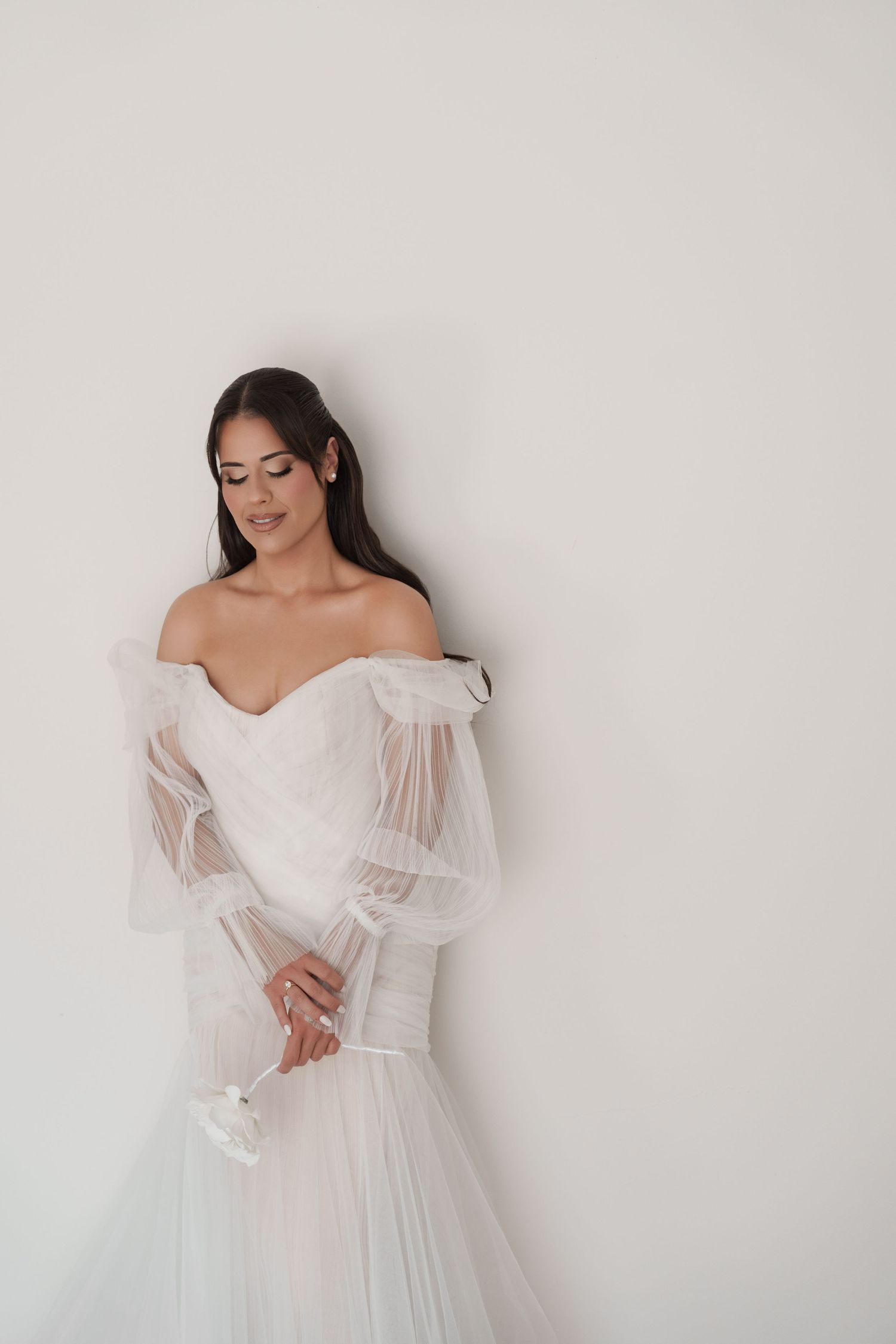 Bride in a flowing off-shoulder tulle wedding dress leaning against a white wall, looking down.
