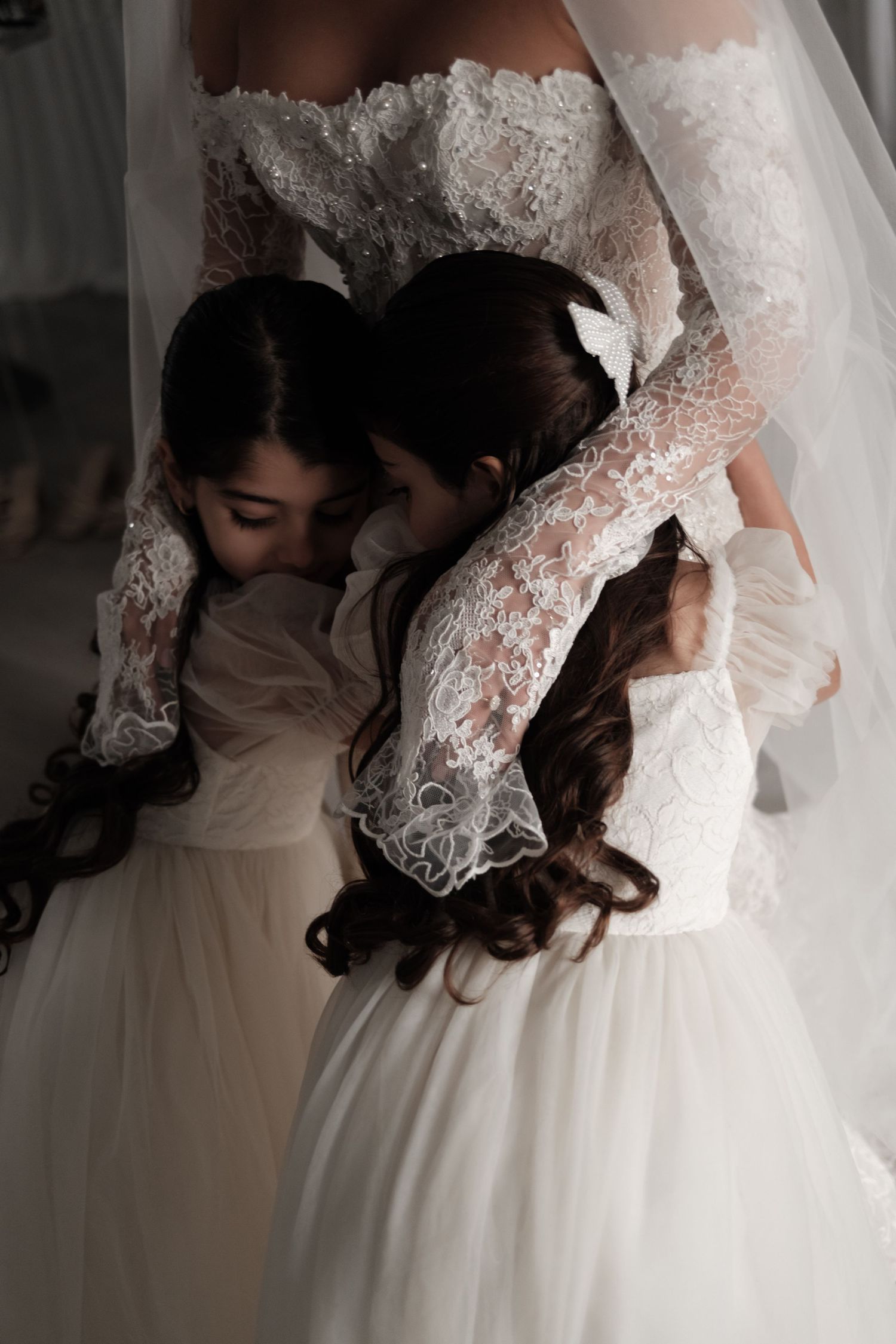 A bride in a lace wedding dress lovingly embraces two young girls in white dresses.