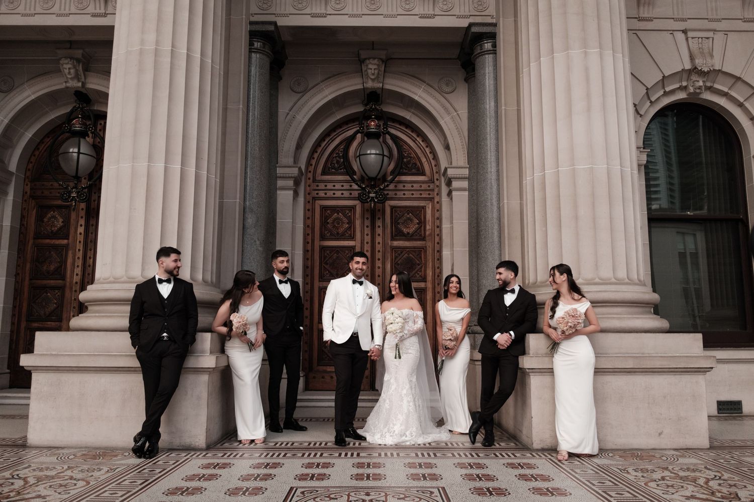 Wedding party posing before grand wooden doors of a classical stone building in elegant attire.
