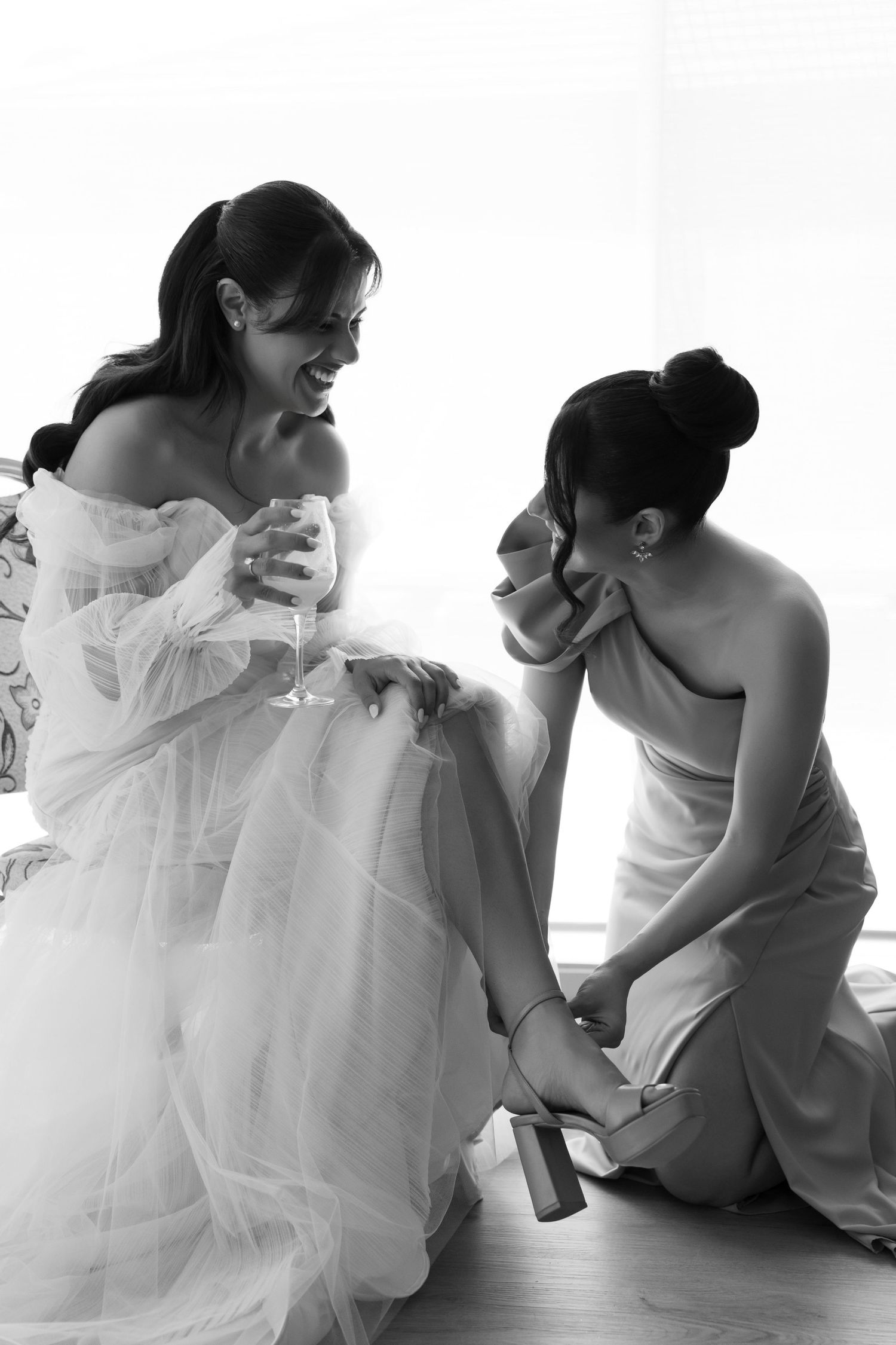 Bridesmaid helps bride with shoes while bride sips champagne in black and white wedding photo.