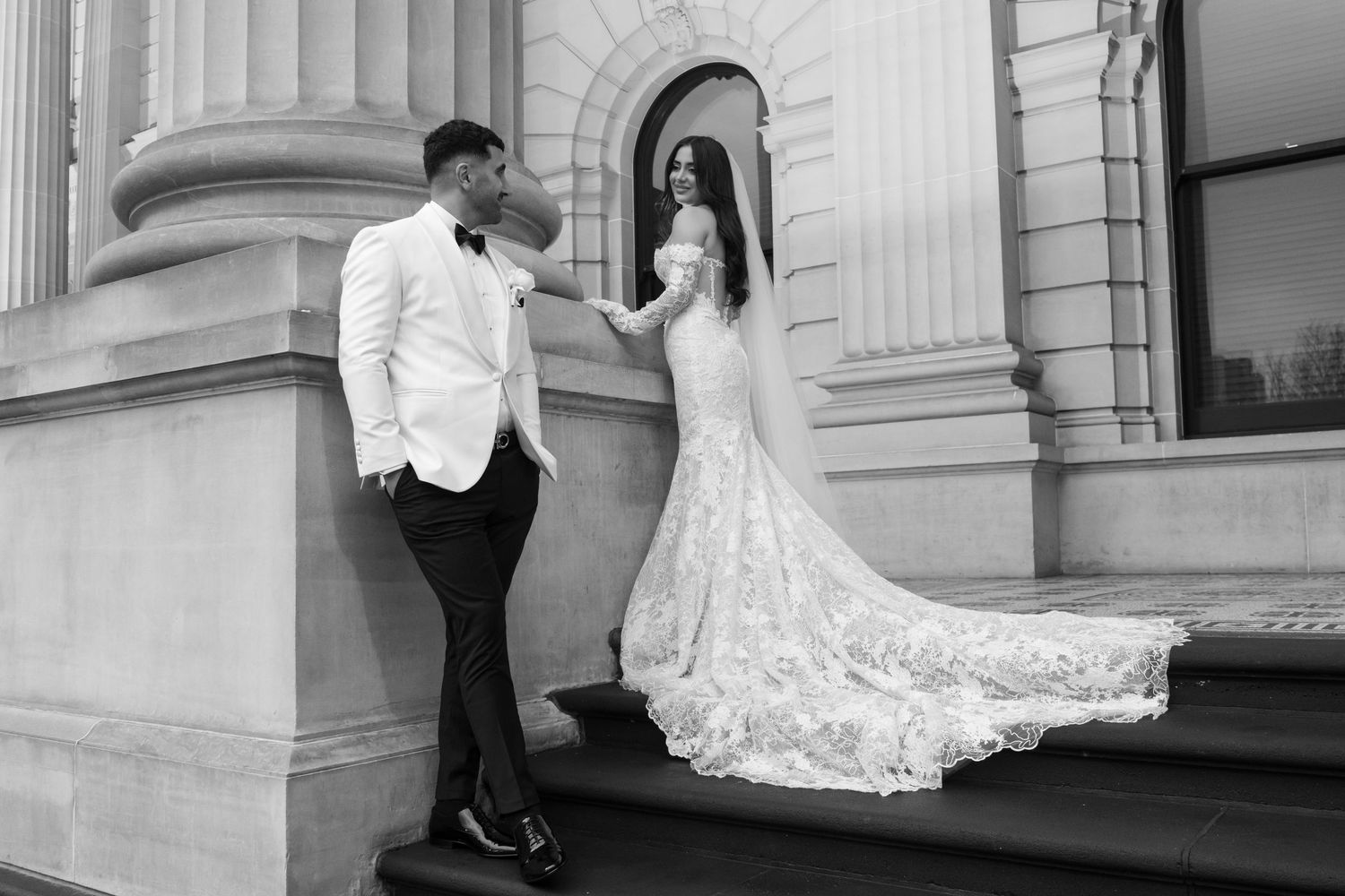 Black and white image of a bride and groom on steps of an ornate building, her lace train flowing.