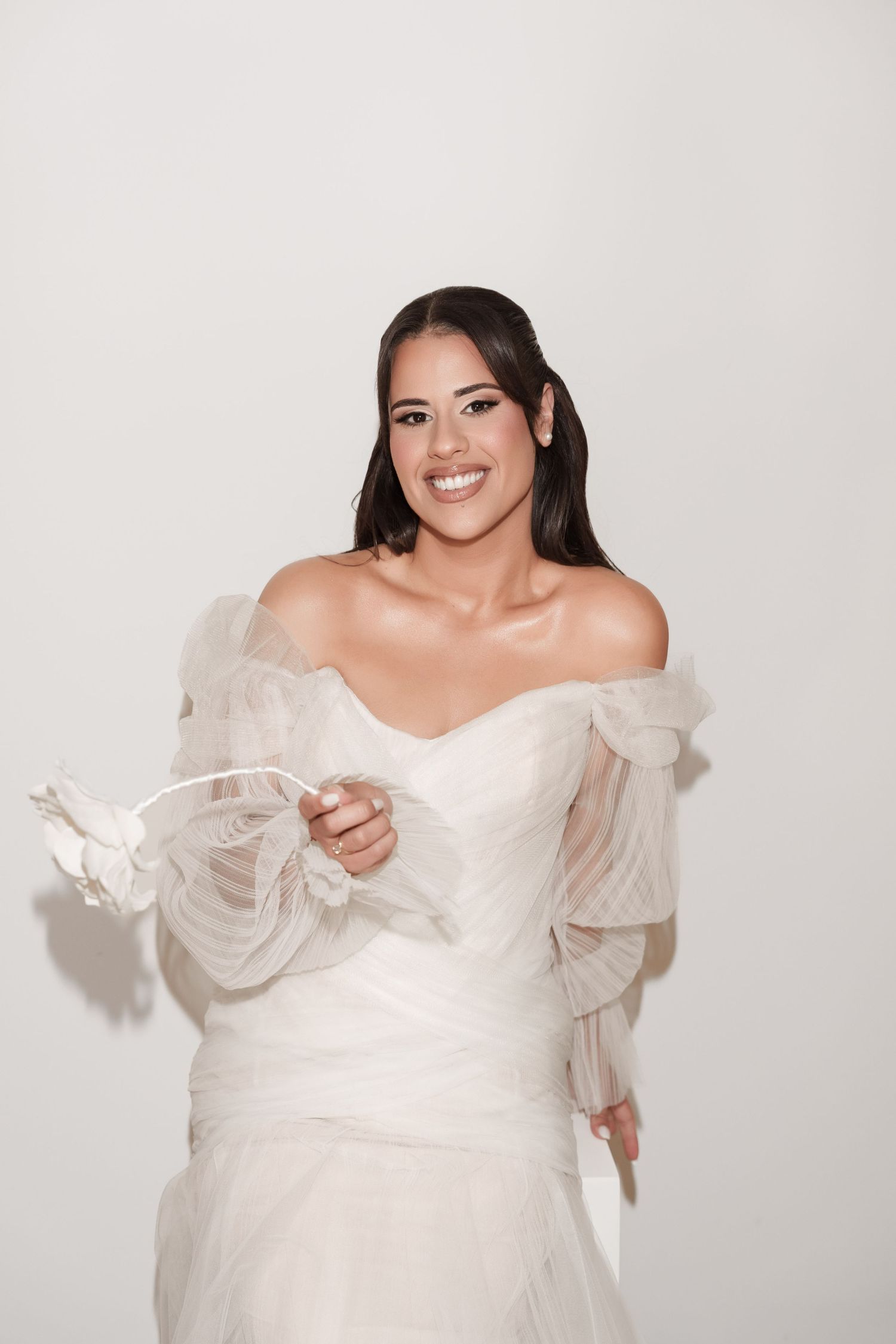 Smiling bride in a white off-shoulder tulle wedding dress holding a delicate fabric flower accessory.