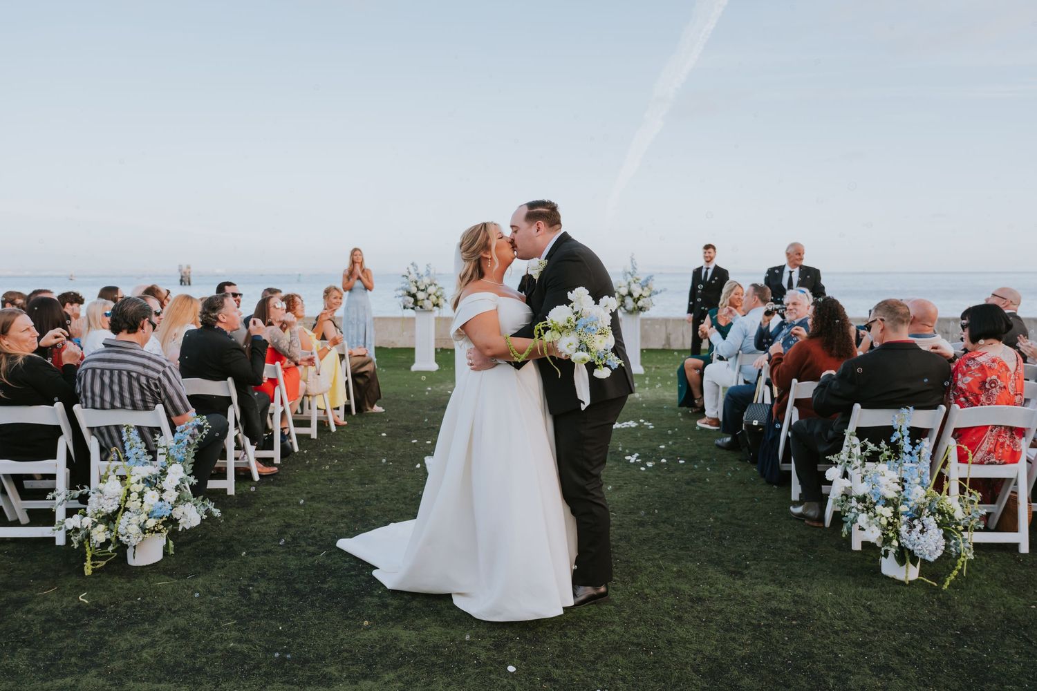 Bride and groom share first kiss at outdoor wedding ceremony on lush green lawn with ocean view backdrop.