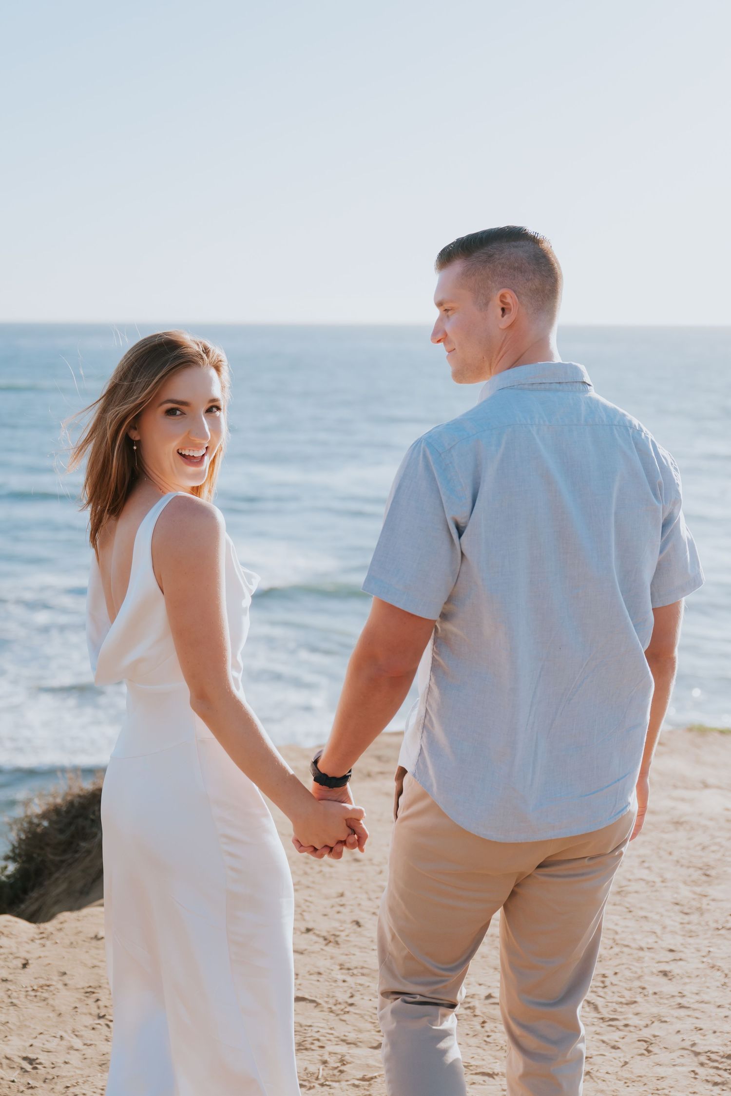 Couple holding hands on sandy beach, woman in white dress smiling over shoulder, ocean backdrop.