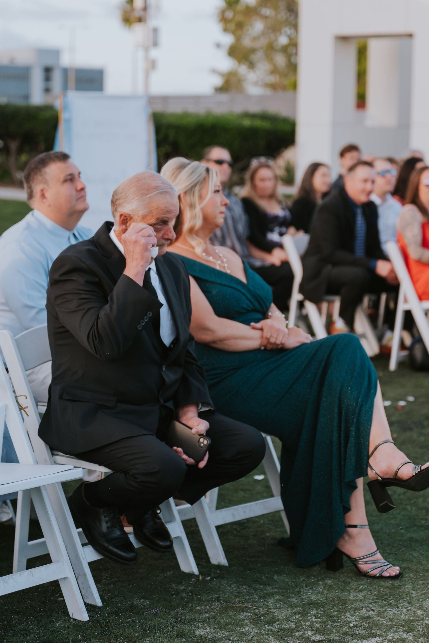 Emotional wedding guest in black suit wiping tears while seated outdoors at a wedding ceremony.