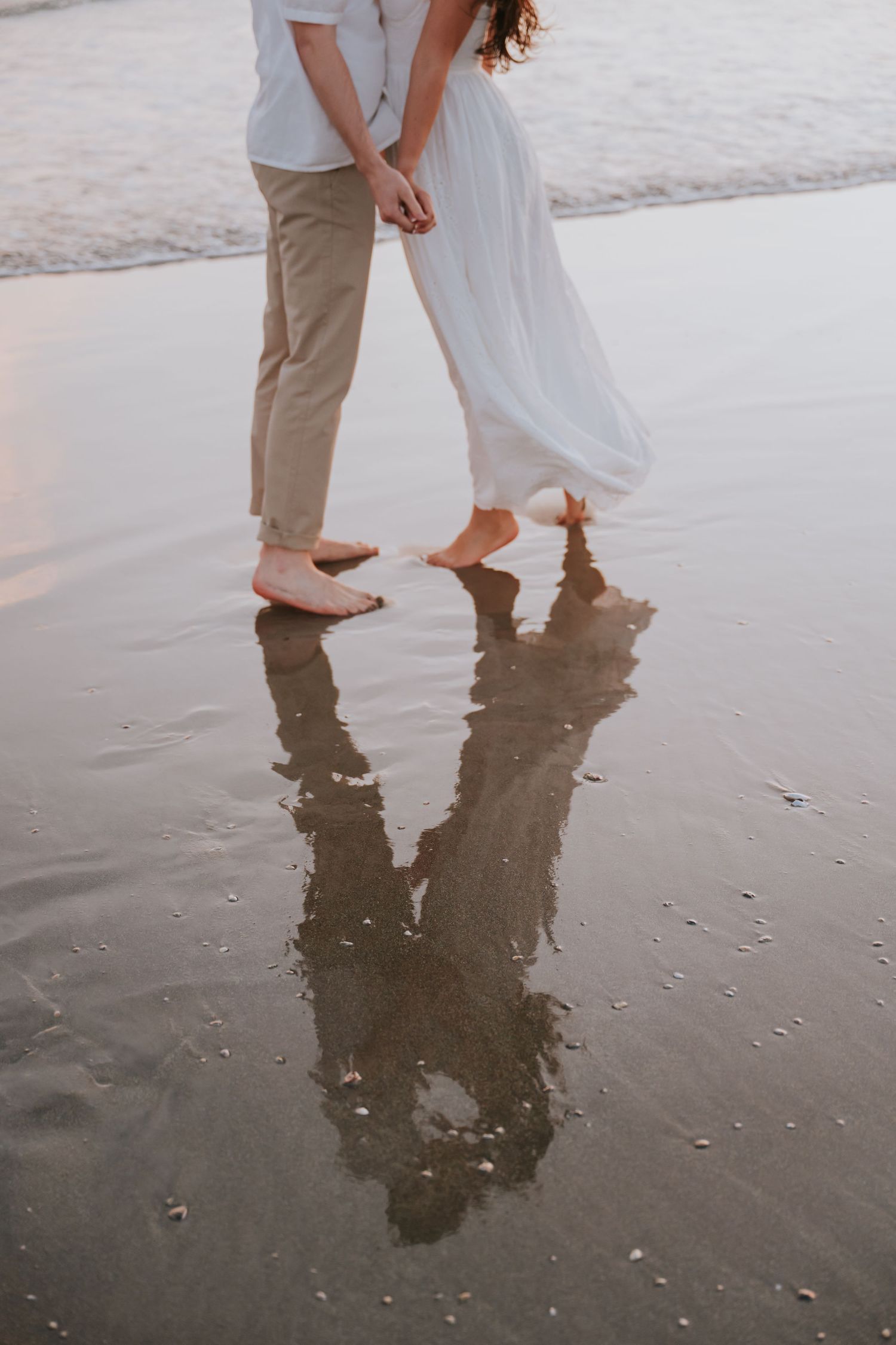 Couple holding hands barefoot on wet beach sand, reflections visible, woman in white dress.