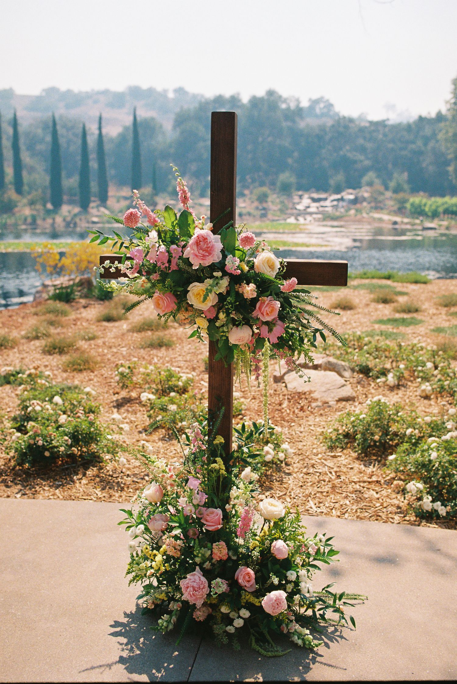 Wooden cross adorned with pink and white floral arrangements at an outdoor wedding ceremony with scenic lake backdrop.