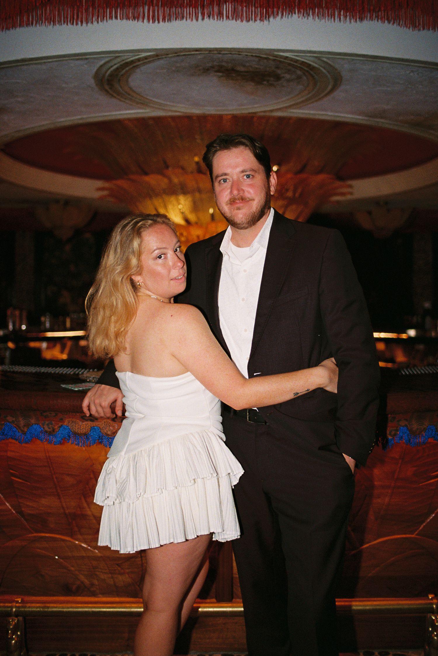Elegant couple posing in formal attire at an ornate venue, woman in white mini dress, man in black suit.