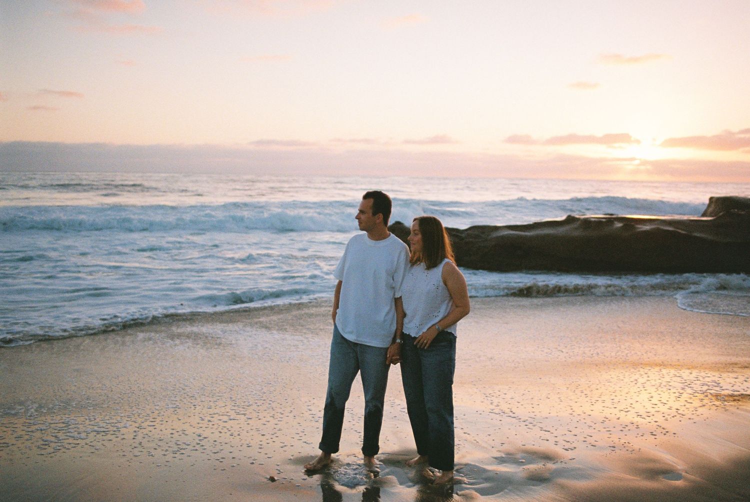 Couple holding hands on sandy beach at sunset with ocean waves and colorful sky in background.