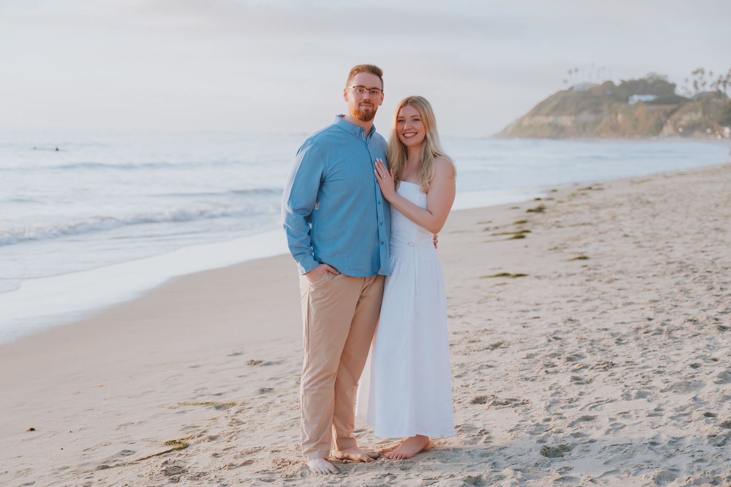 Couple posing together on a sandy beach at sunset, man in blue shirt and woman in white dress.