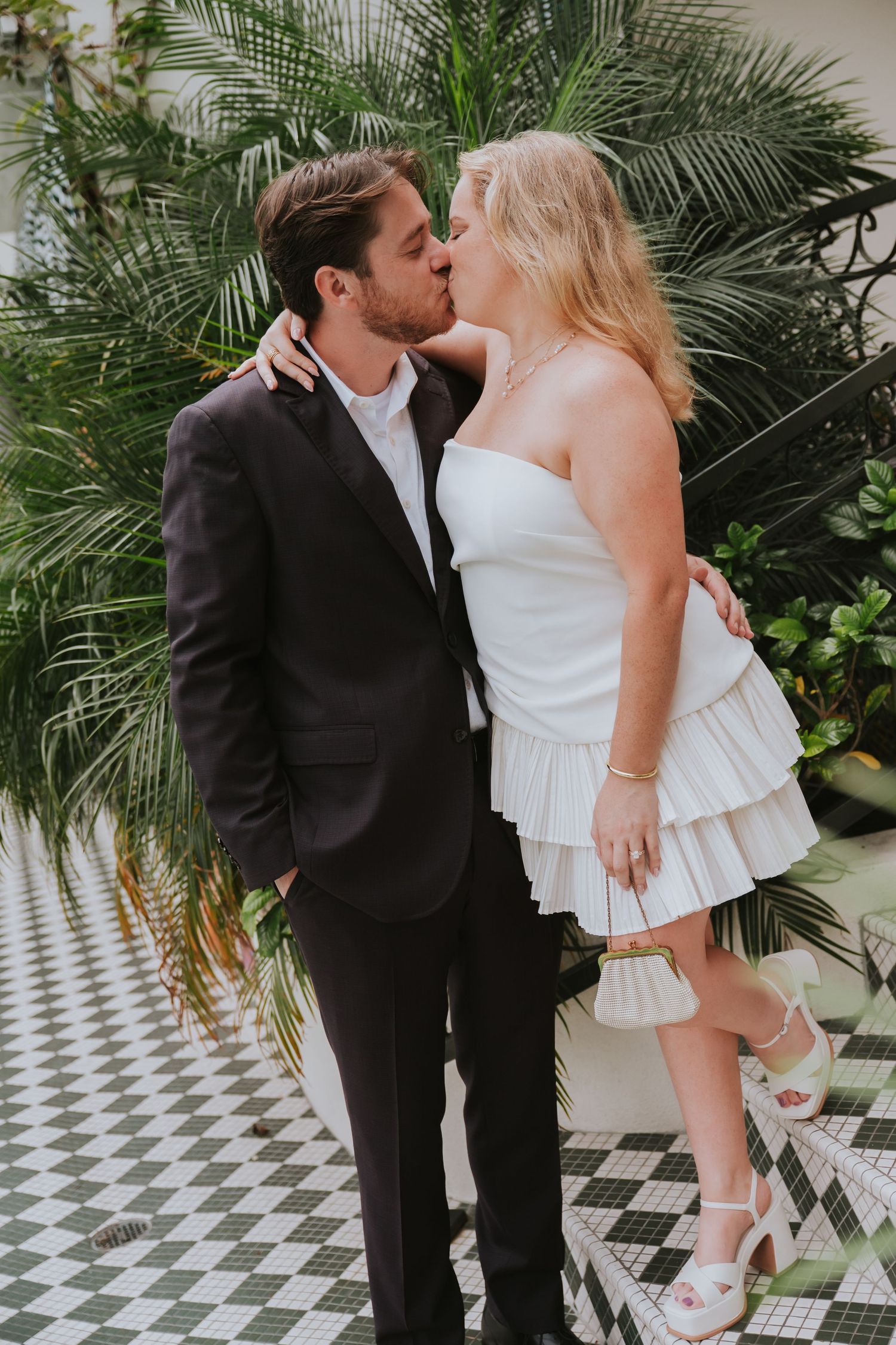 Newlywed couple sharing a romantic kiss in a tropical garden setting, bride in white mini dress.