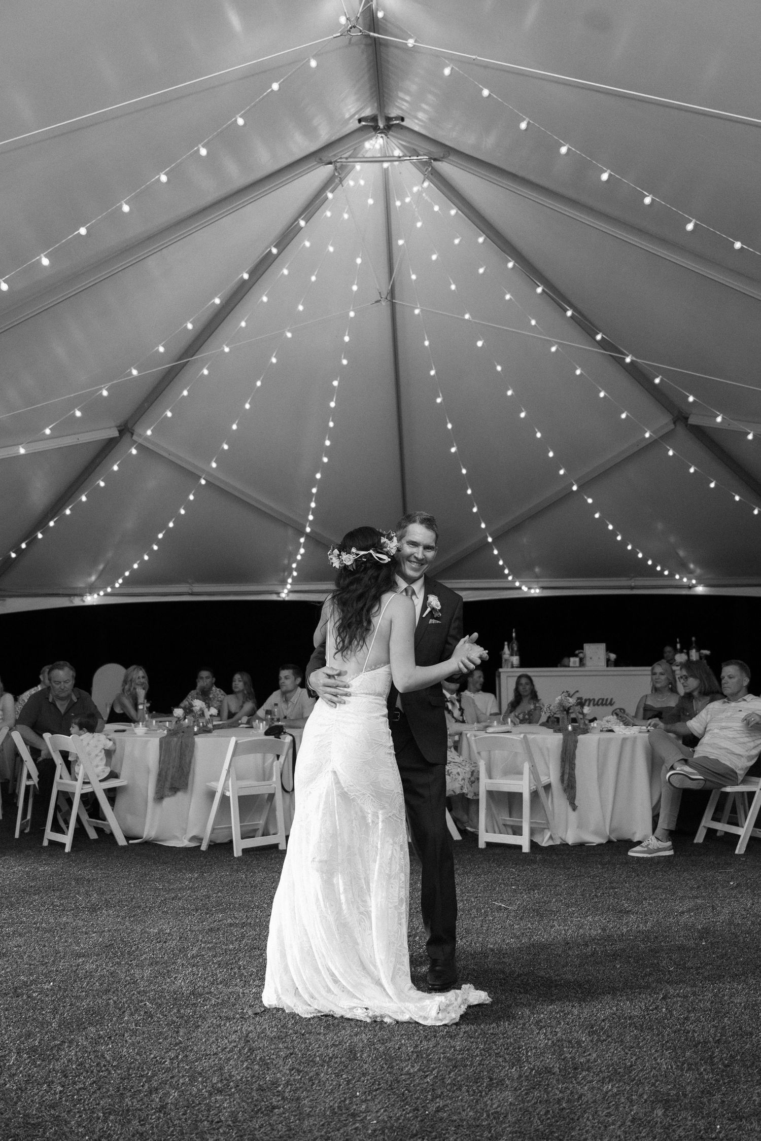 A couple shares their first dance under a white tent decorated with twinkling string lights at an outdoor wedding reception.