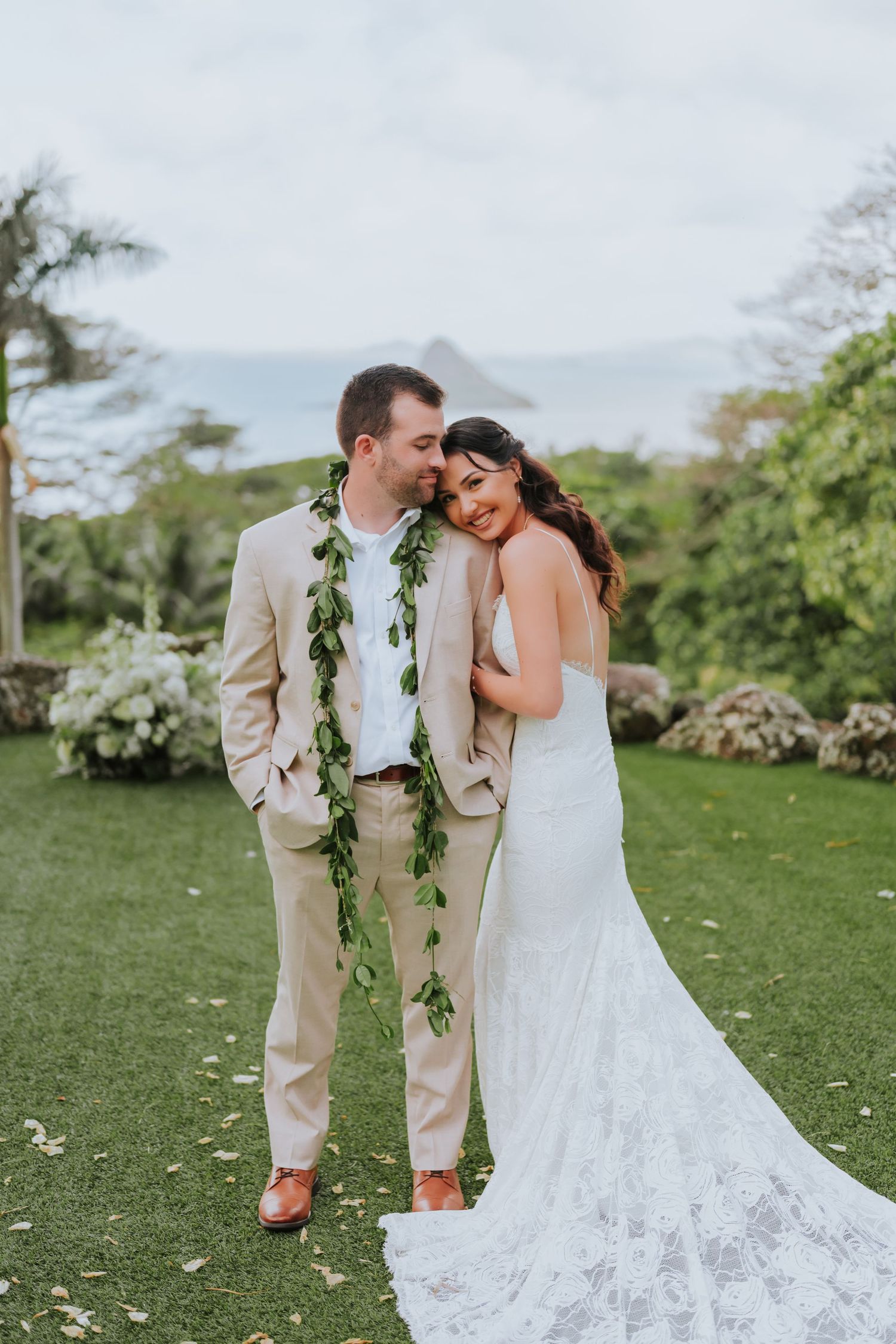 Newlyweds share an intimate moment on a grassy hillside with white floral decorations and greenery.