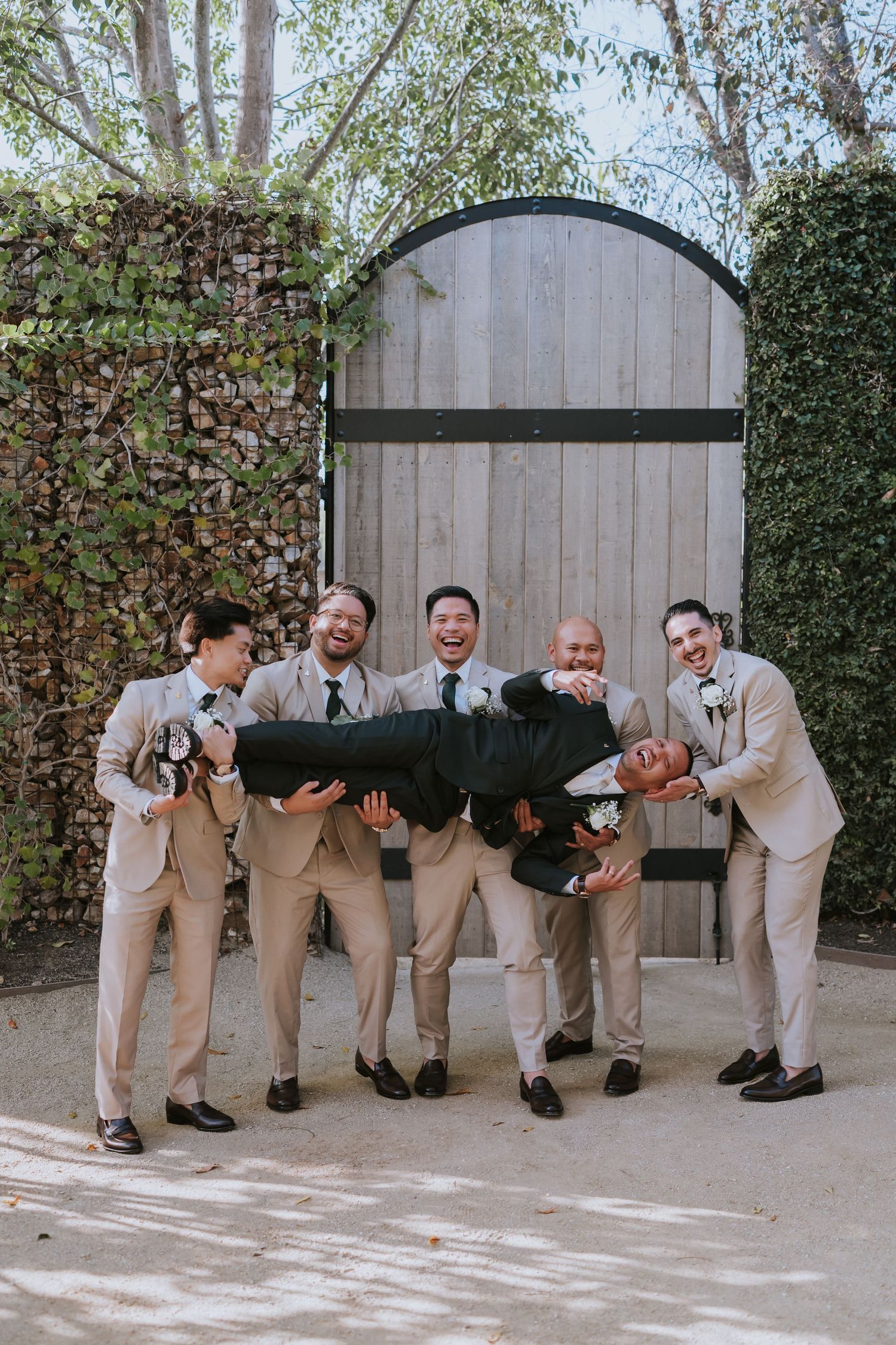 A playful wedding party in tan suits poses in a sequence showing them falling like dominos against a wooden fence.