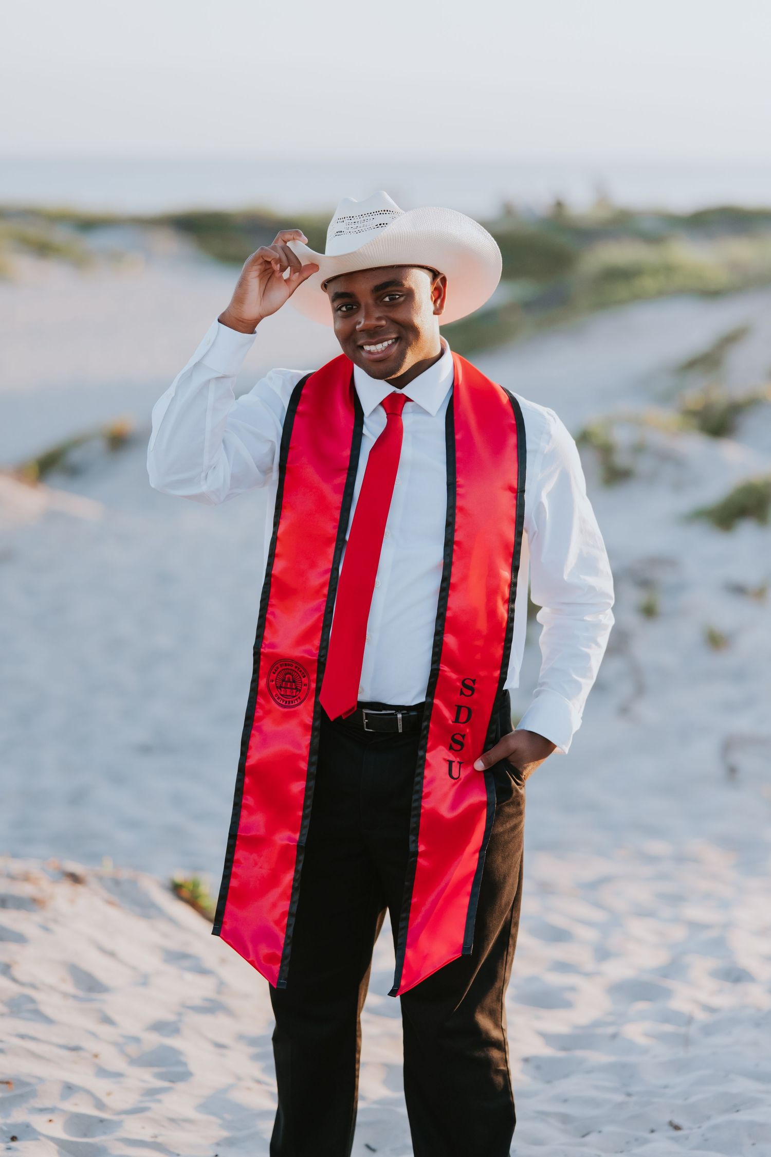 Smiling graduate in white cowboy hat, red sash, and tie poses on sandy beach dunes in formal attire.