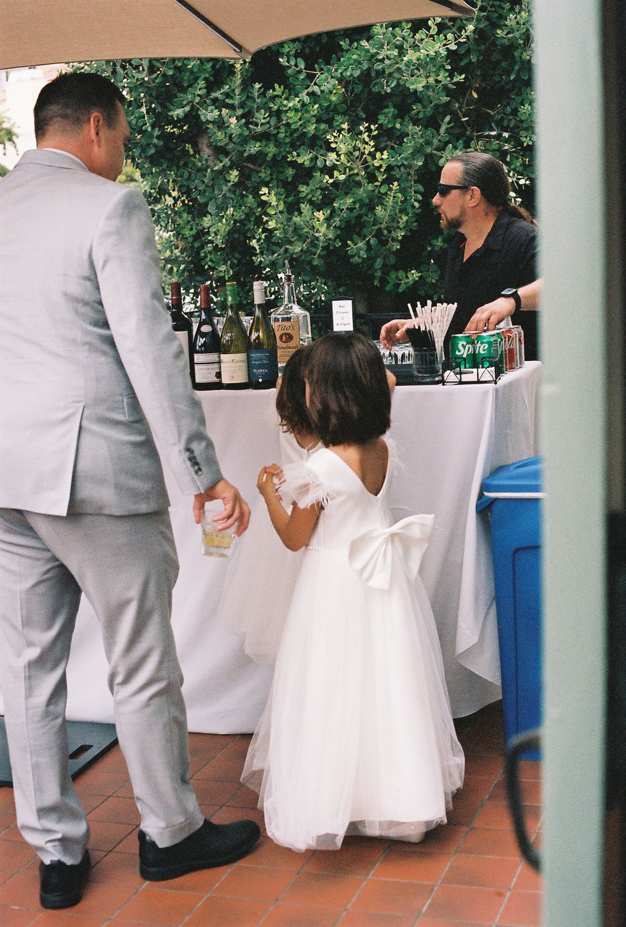 A flower girl in a white tulle dress with bow stands at an outdoor wedding bar next to a man in a gray suit.