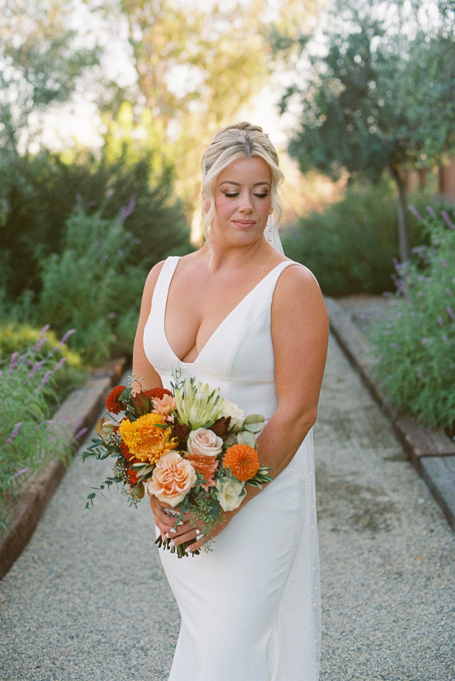Bride in white gown holding orange and peach bouquet stands on garden path in warm golden light.