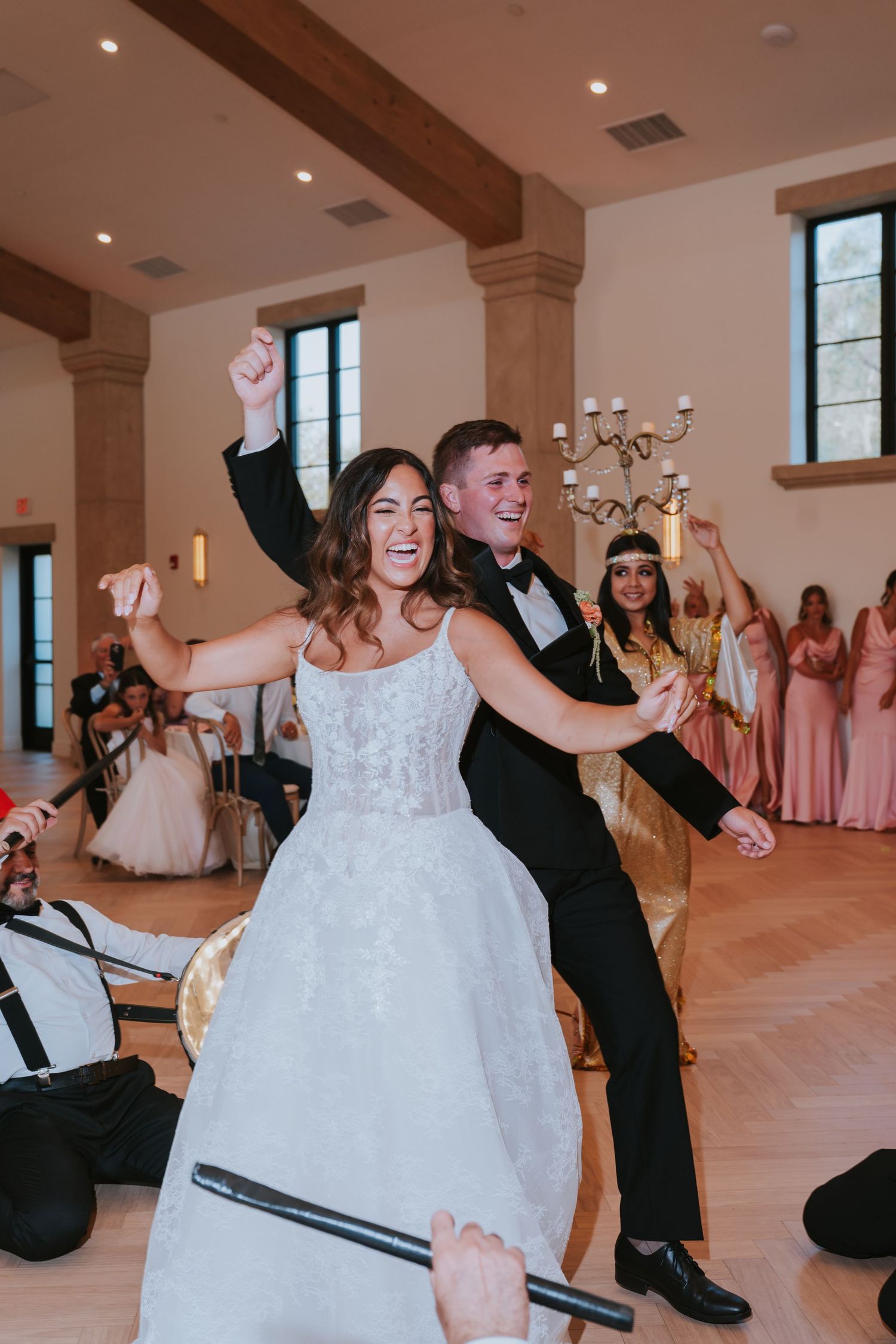 Joyful bride and groom dancing at wedding reception, bride in lace gown, groom in tuxedo, guests in pink dresses behind them.