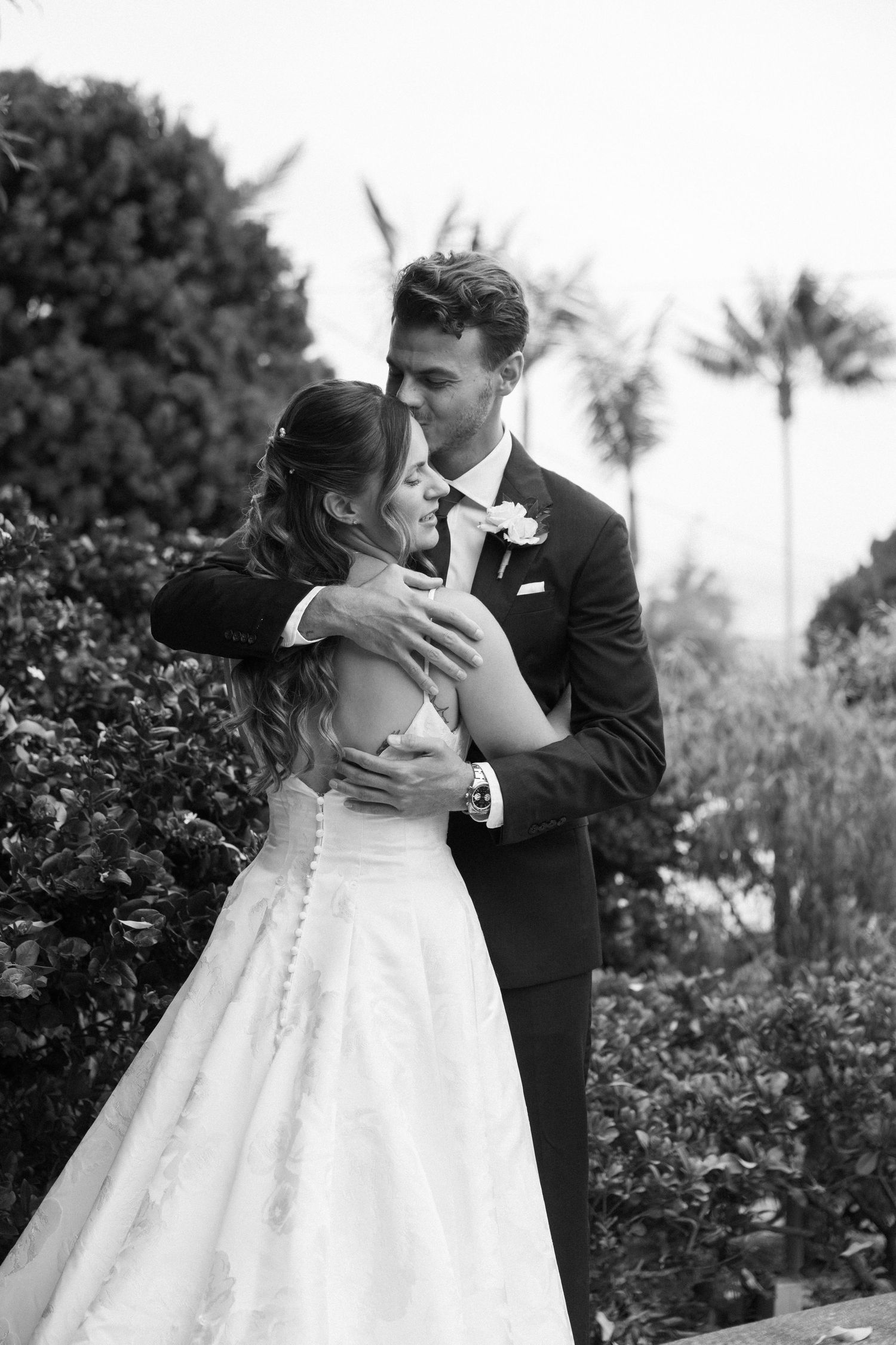 A romantic black and white wedding photo shows a couple embracing in a garden with palm trees in the background.