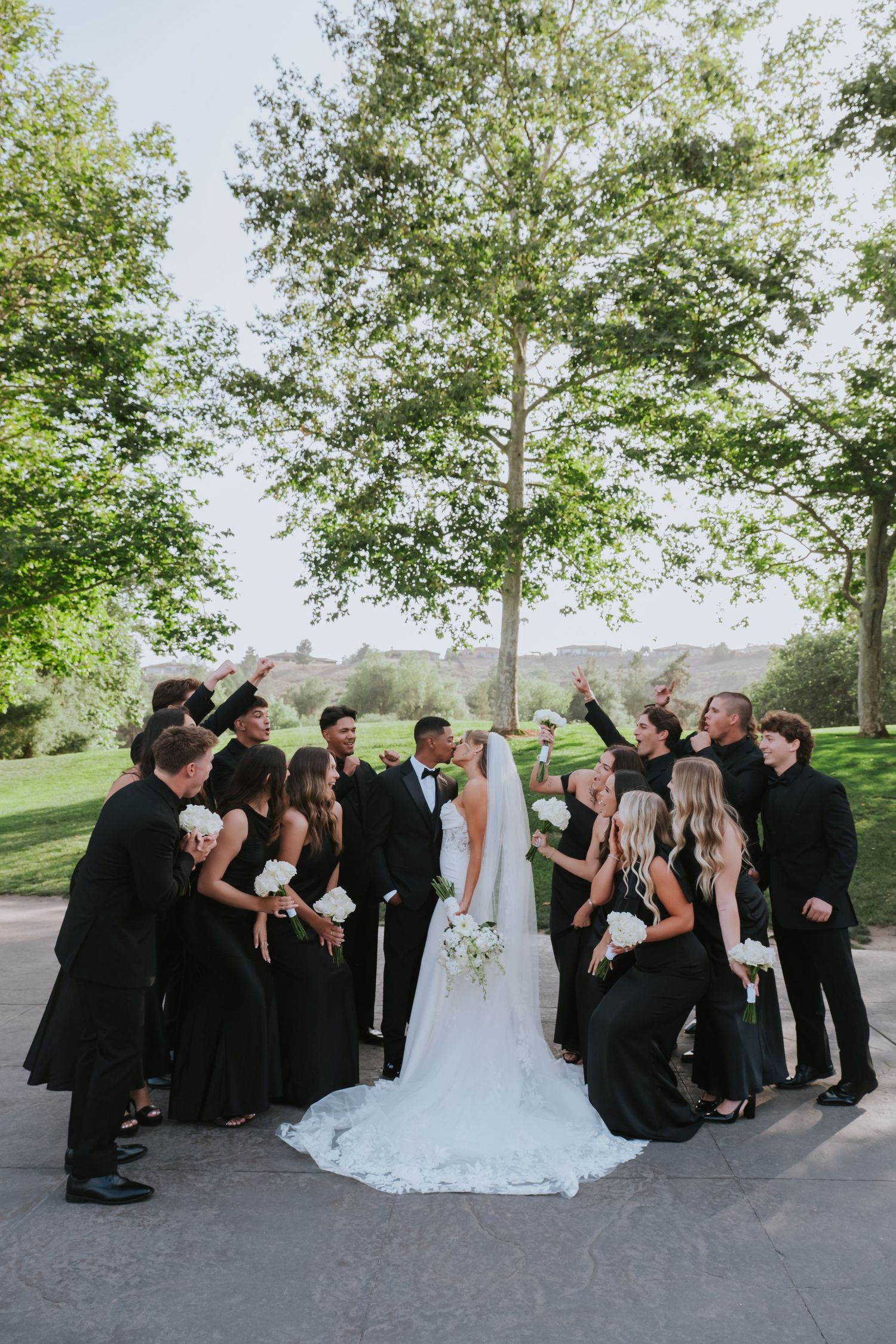 Wedding party in black attire celebrating with bride and groom outdoors among trees.