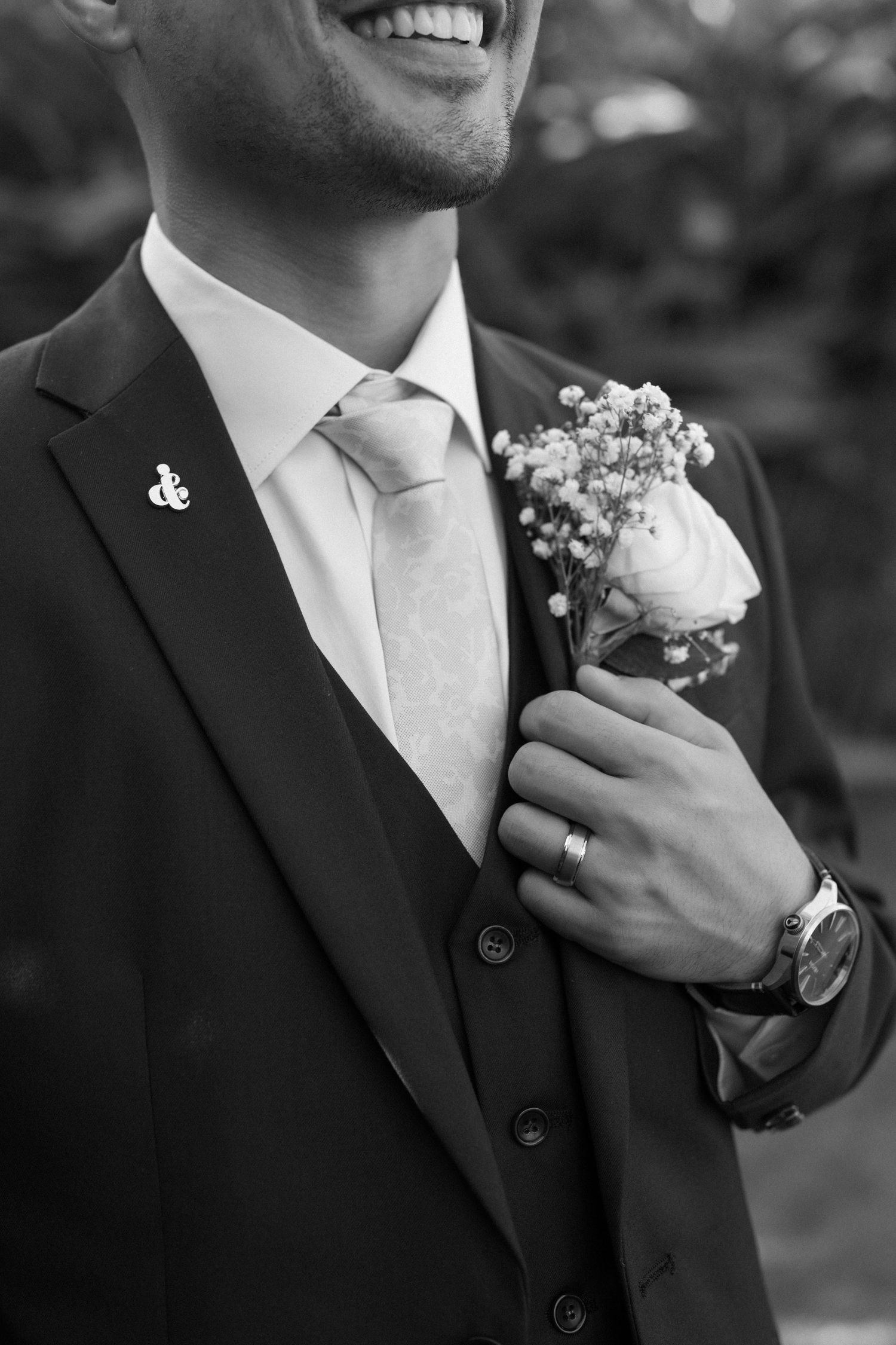 A close-up black and white photo of a groom in a three-piece suit with boutonniere and wedding ring.