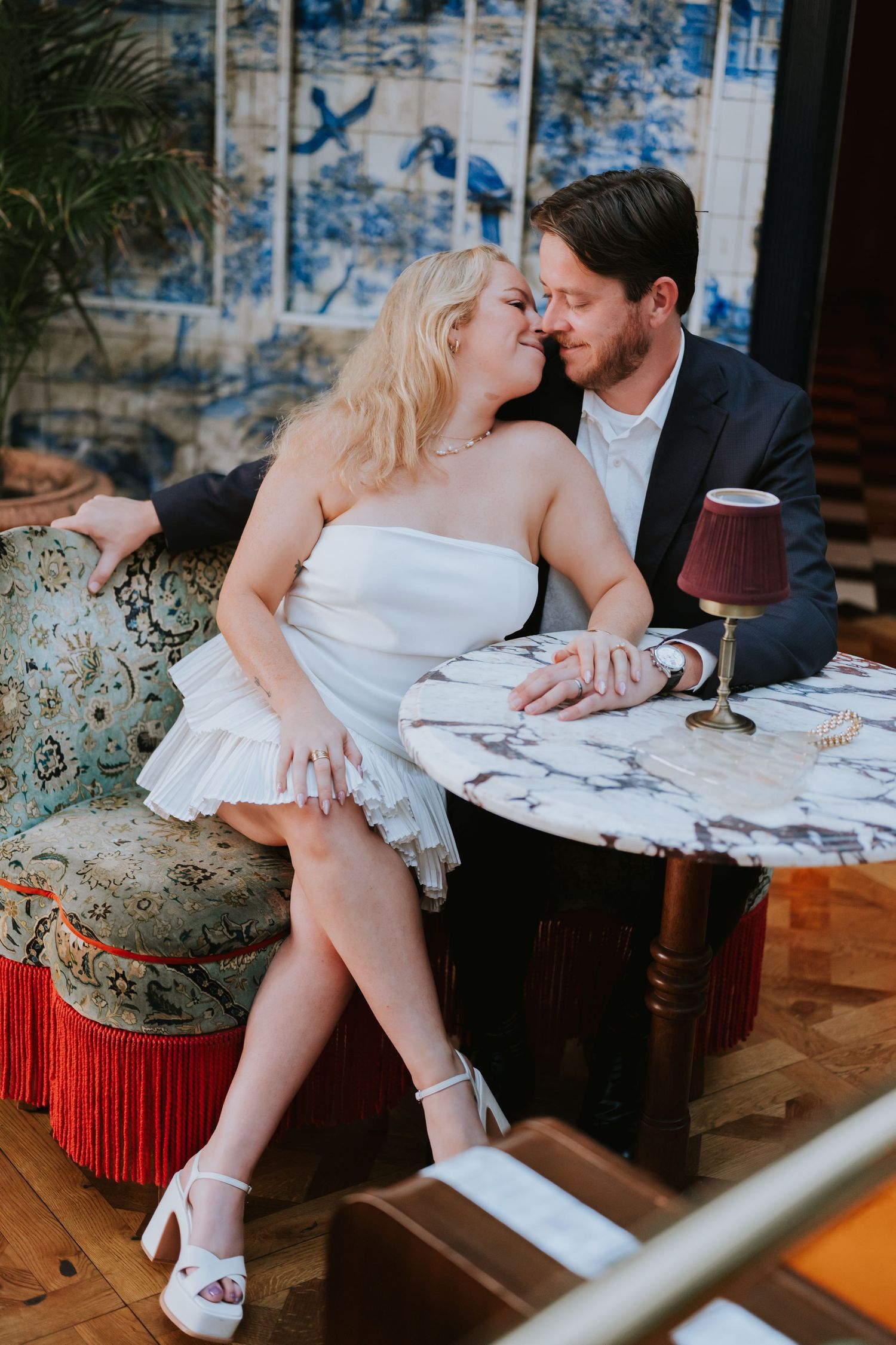 Bride and groom sit closely together at a marble table on a vintage sofa, nearly kissing.