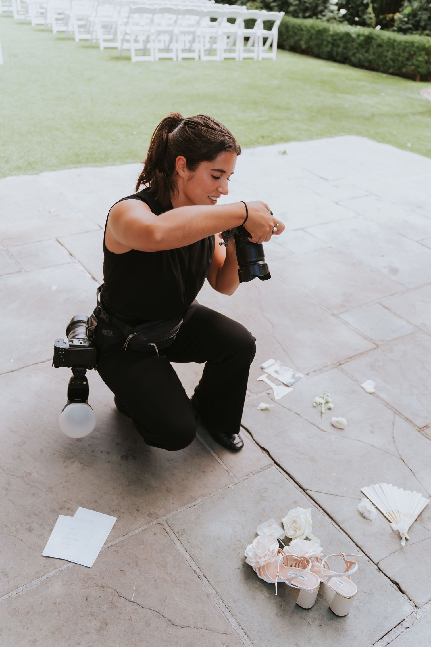 Female photographer kneeling on stone patio, photographing wedding shoes and accessories with white chairs in background.