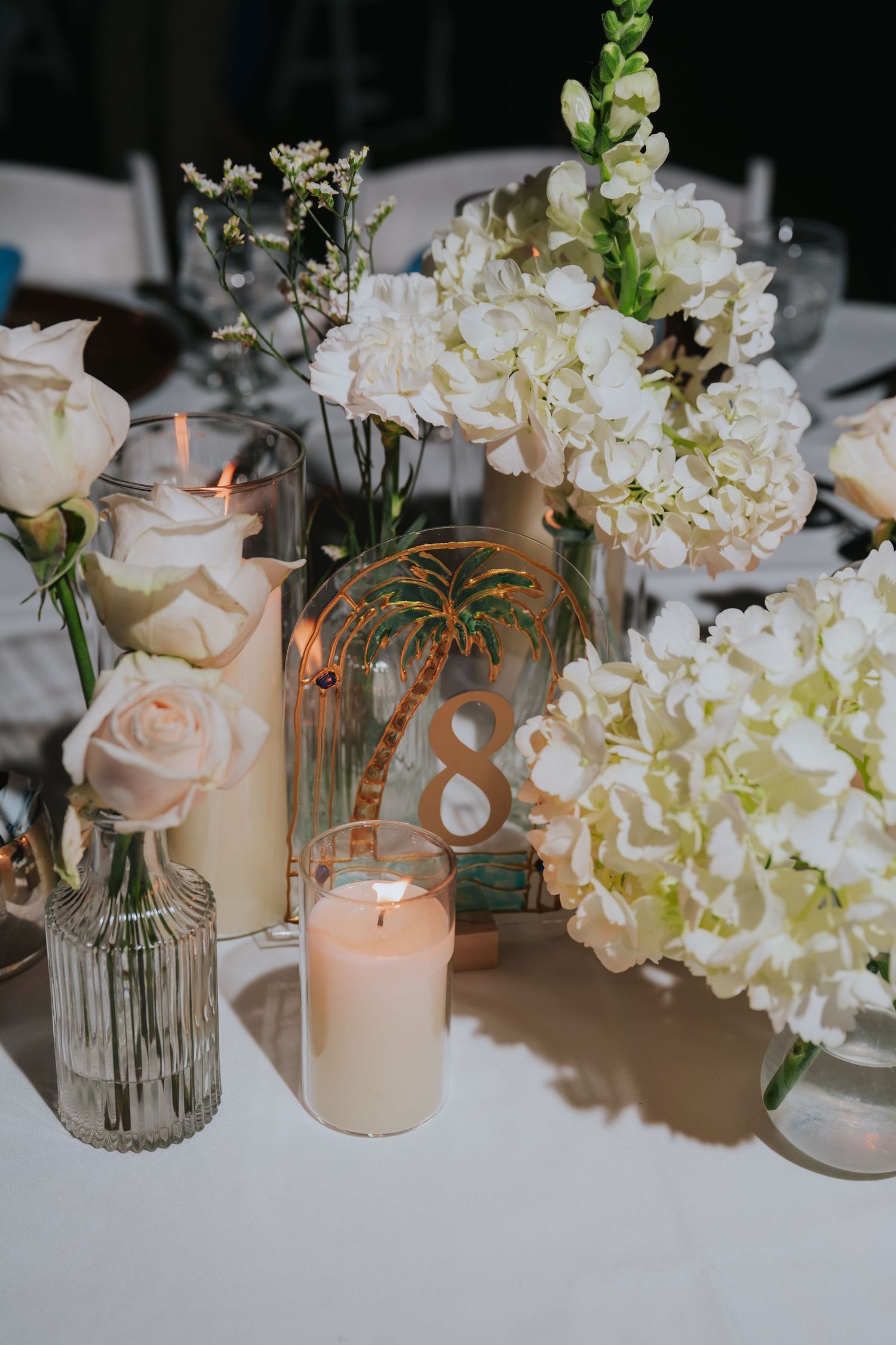 Elegant wedding table centerpiece with white hydrangeas, roses, candles, and palm tree table number.