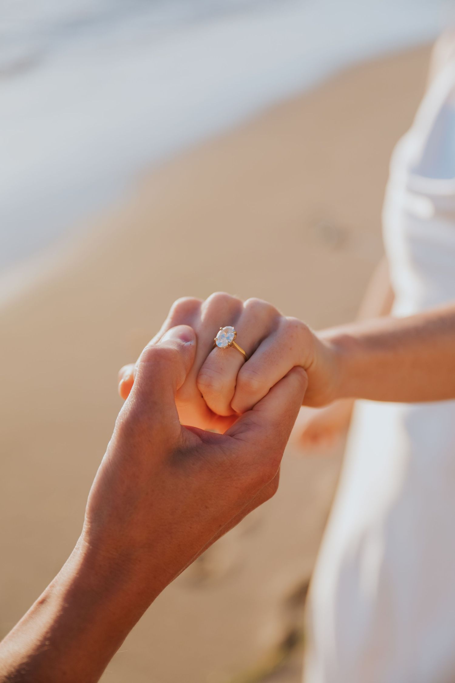 Close-up of a couple holding hands on a beach, showcasing a stunning diamond engagement ring on the woman's finger.