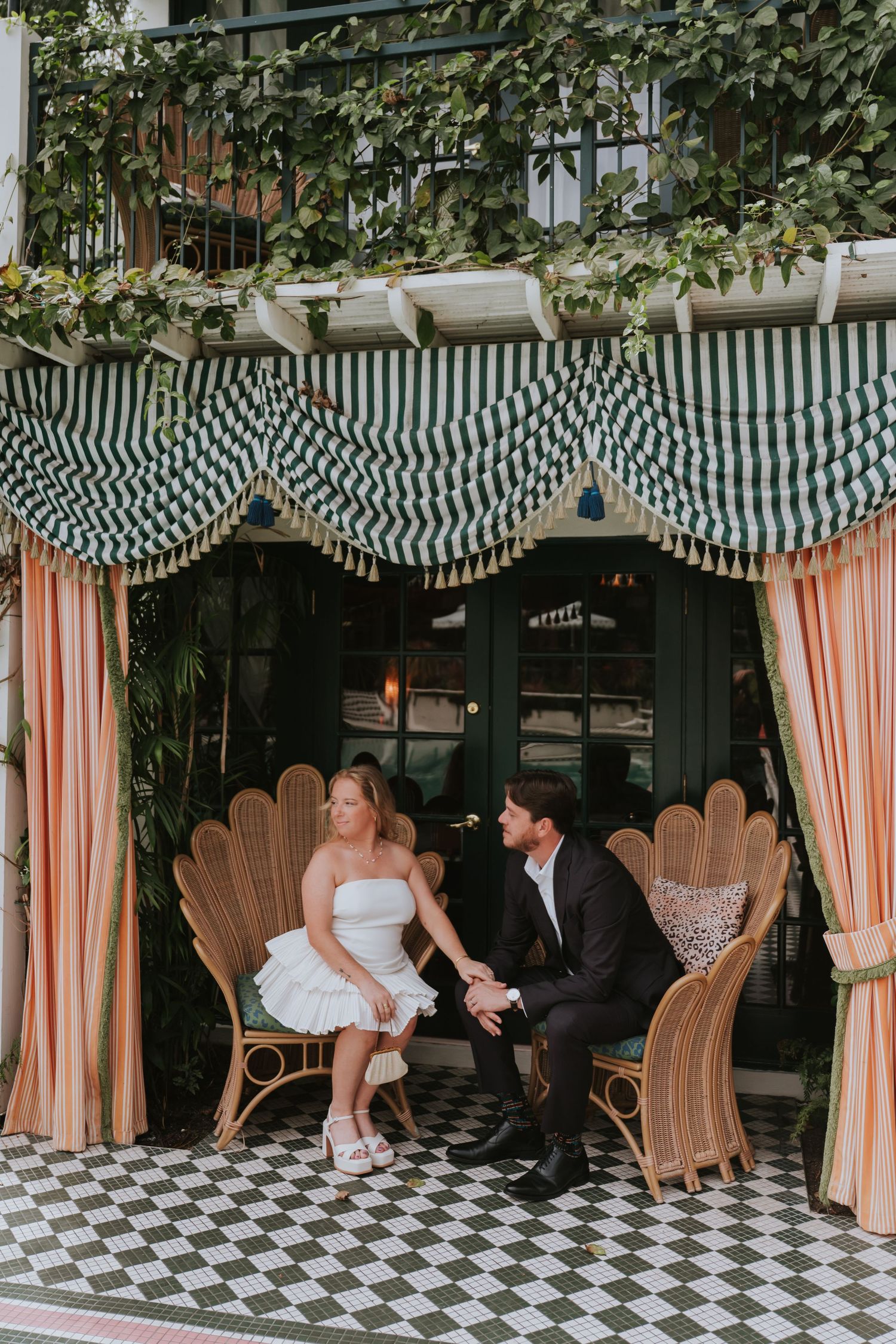 Newlywed couple sitting in rattan shell chairs under striped awning at a charming outdoor venue.