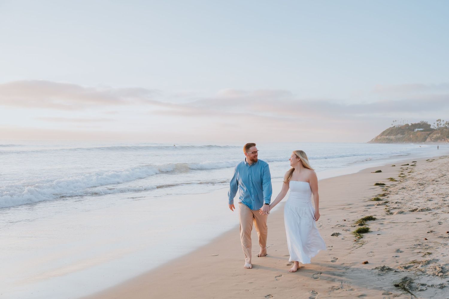 Couple walking hand in hand on a sandy beach at sunset, man in blue shirt and woman in white dress.