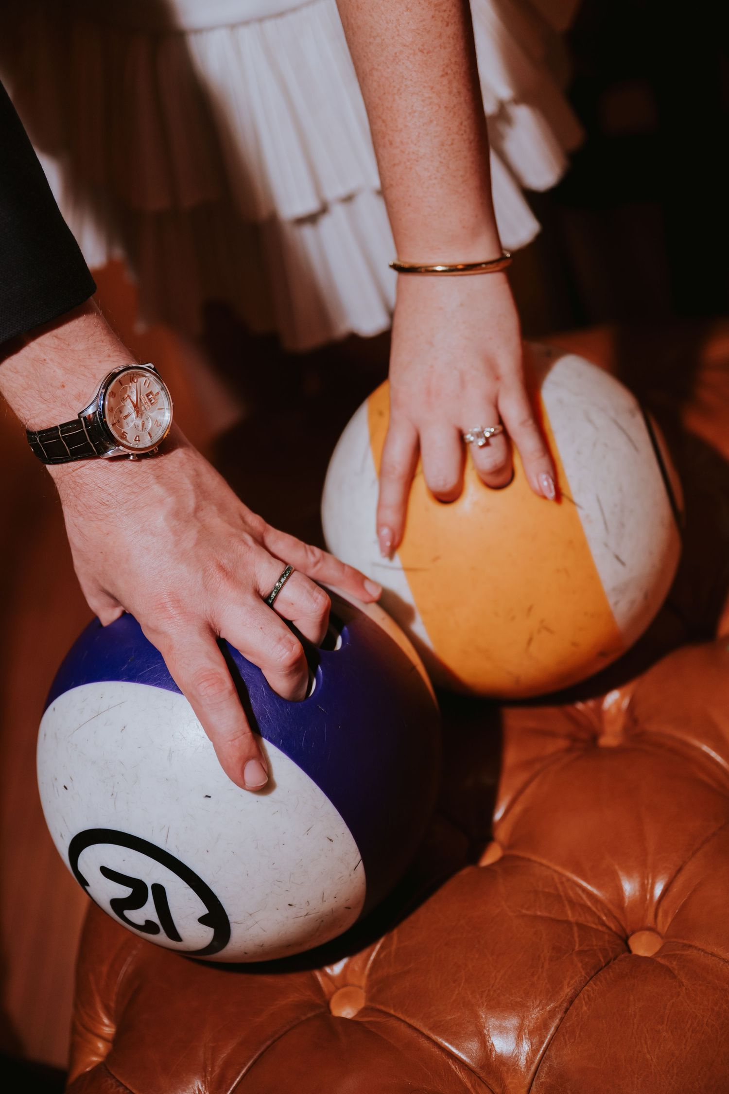 Two hands resting on colorful bingo balls on a leather surface, one wearing a watch and ring, the other a bracelet and ring.