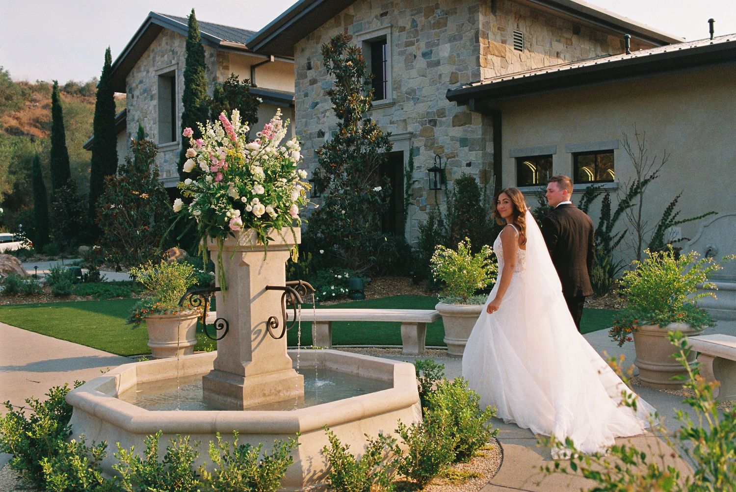 Bride and groom share a romantic moment by a stone fountain outside an elegant venue surrounded by lush gardens.