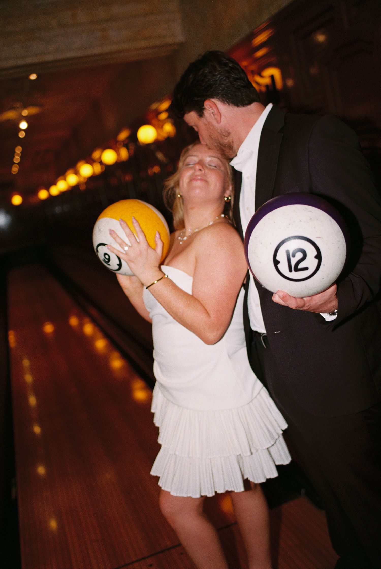 Bride and groom kissing while holding bowling balls on a bowling alley lane at their wedding.