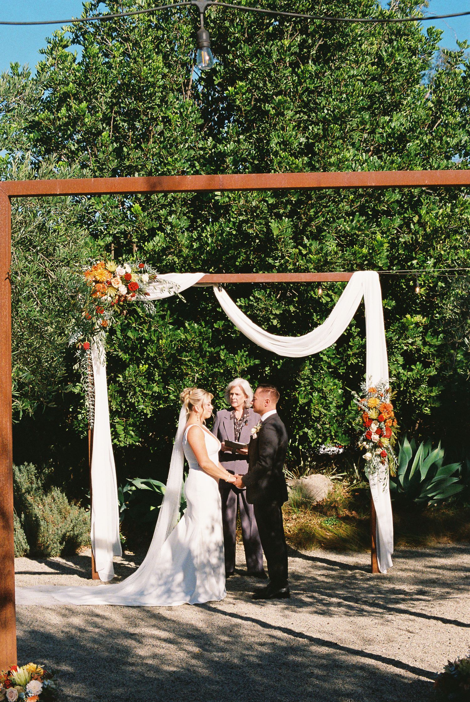 Bride and groom exchange vows under a wooden arch draped with white fabric and fall florals in an outdoor garden ceremony.