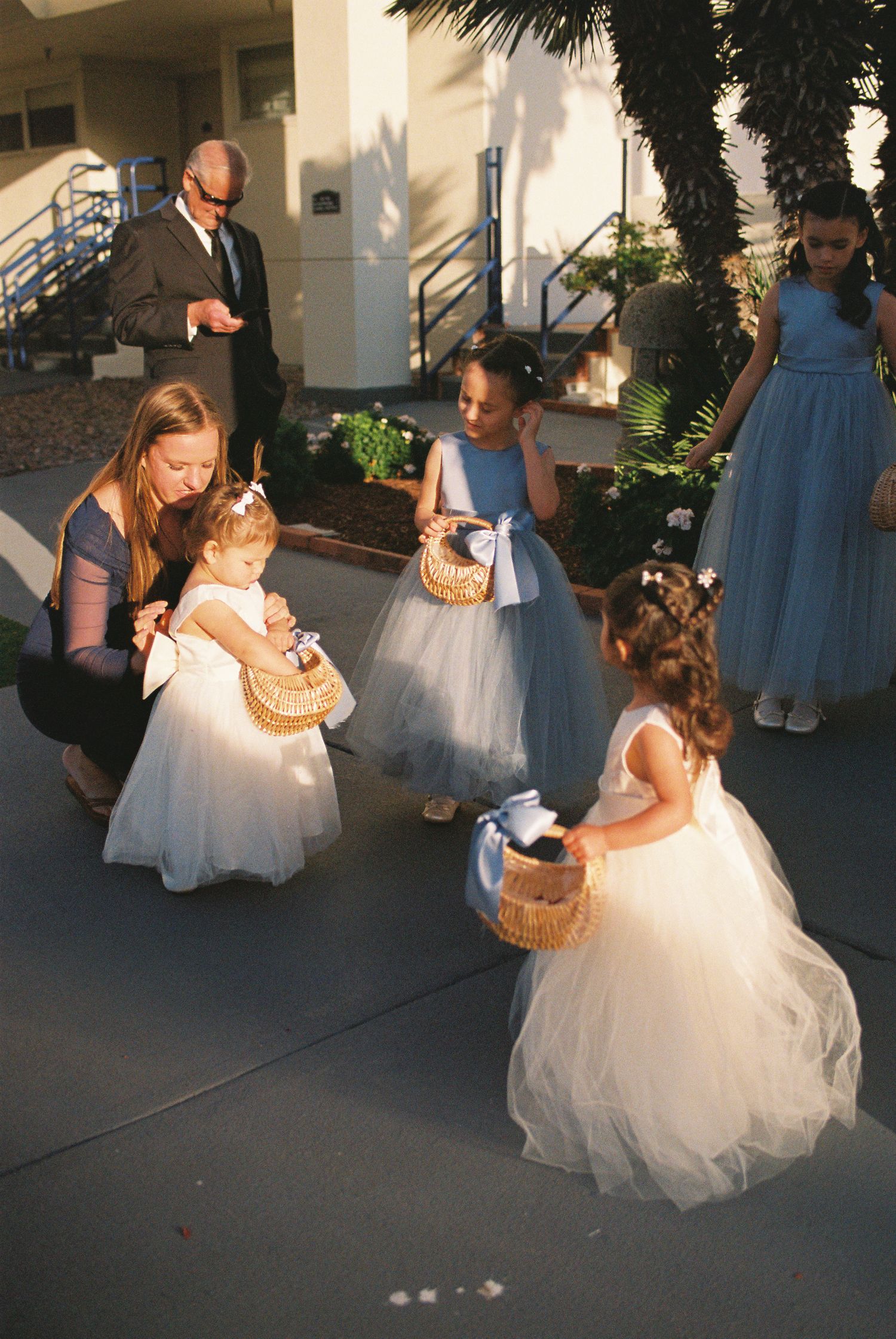 Flower girls in white tulle dresses holding wicker baskets gather outside a wedding venue with an adult woman.