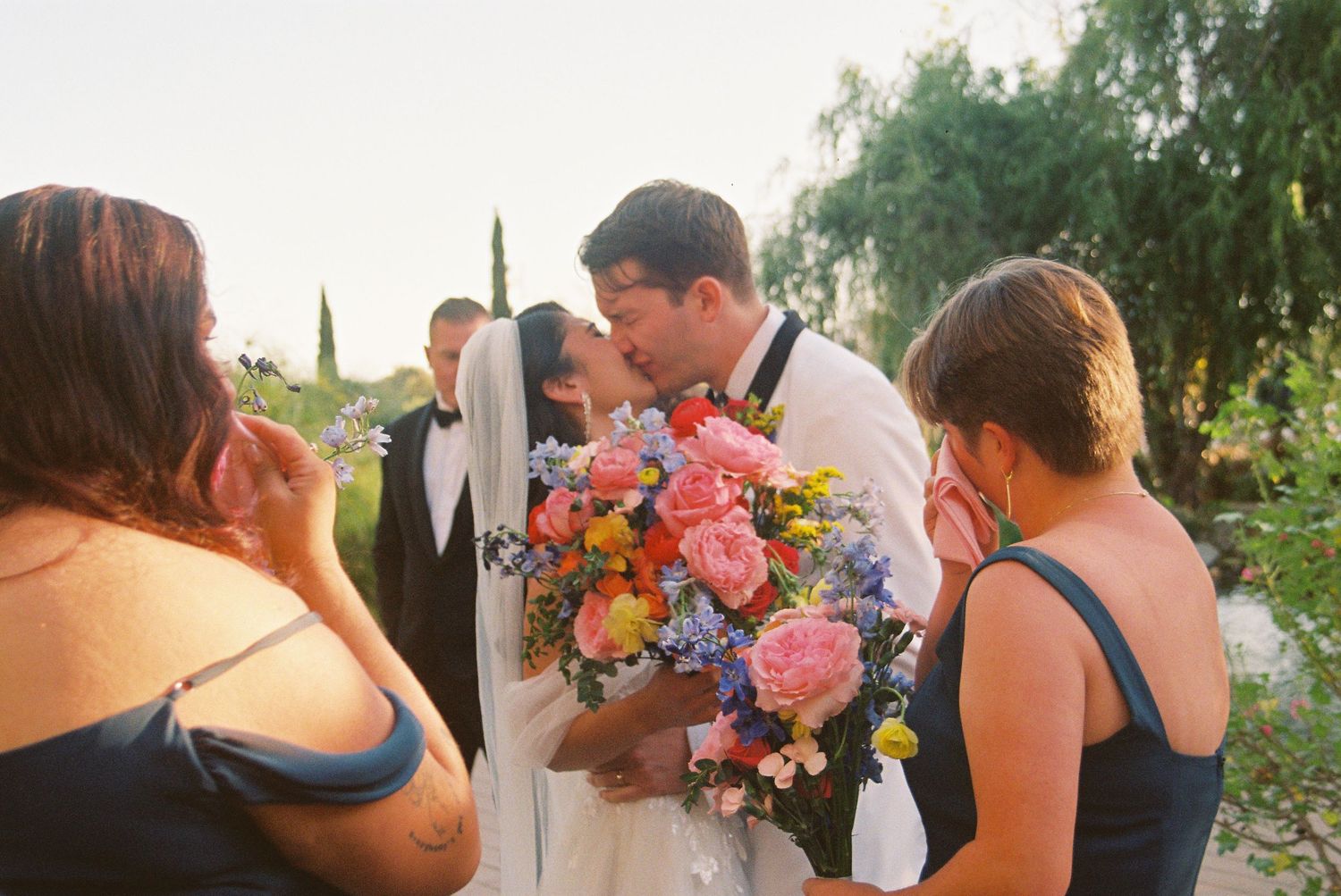 Bride and groom share a kiss at outdoor wedding ceremony, surrounded by bridesmaids holding colorful bouquet.