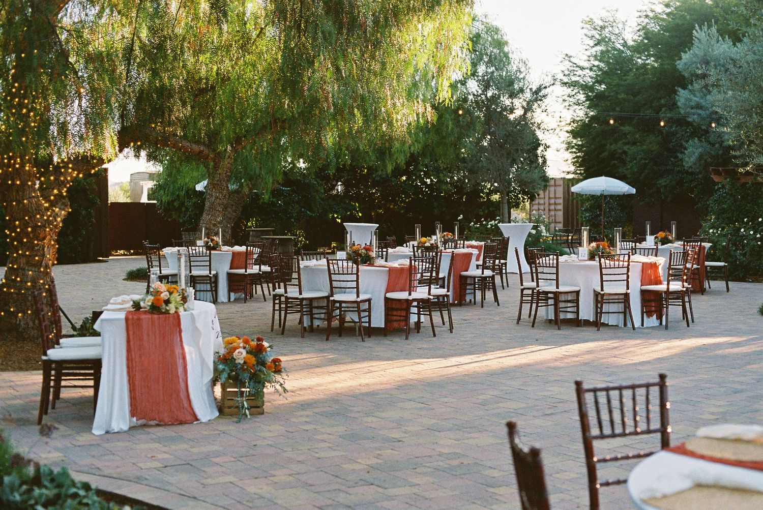 Elegant outdoor wedding reception with round tables, terracotta linens, floral centerpieces, and string lights on a patio.