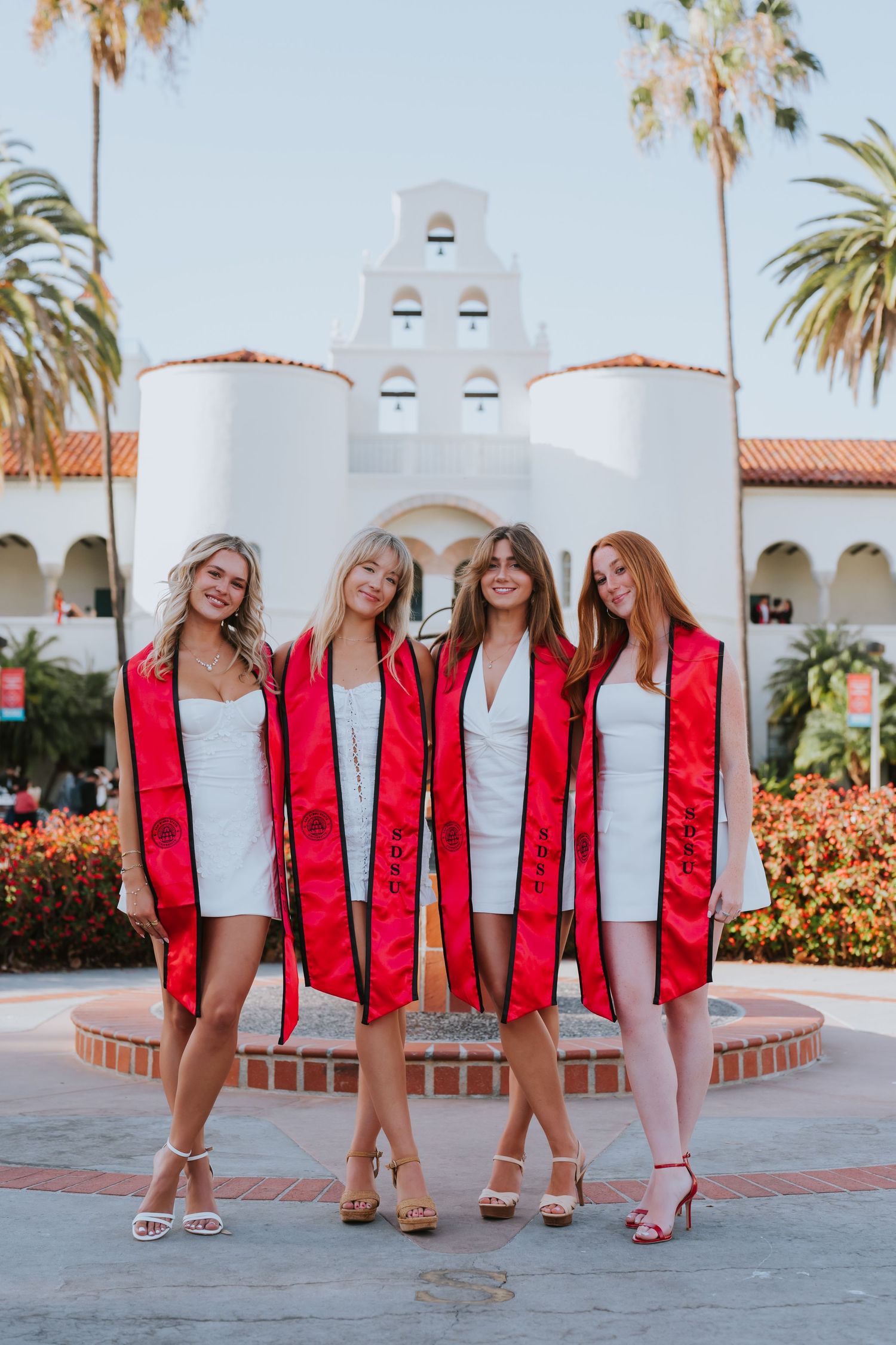 Four female graduates in white dresses and red sashes pose in front of a white bell tower at SDSU.