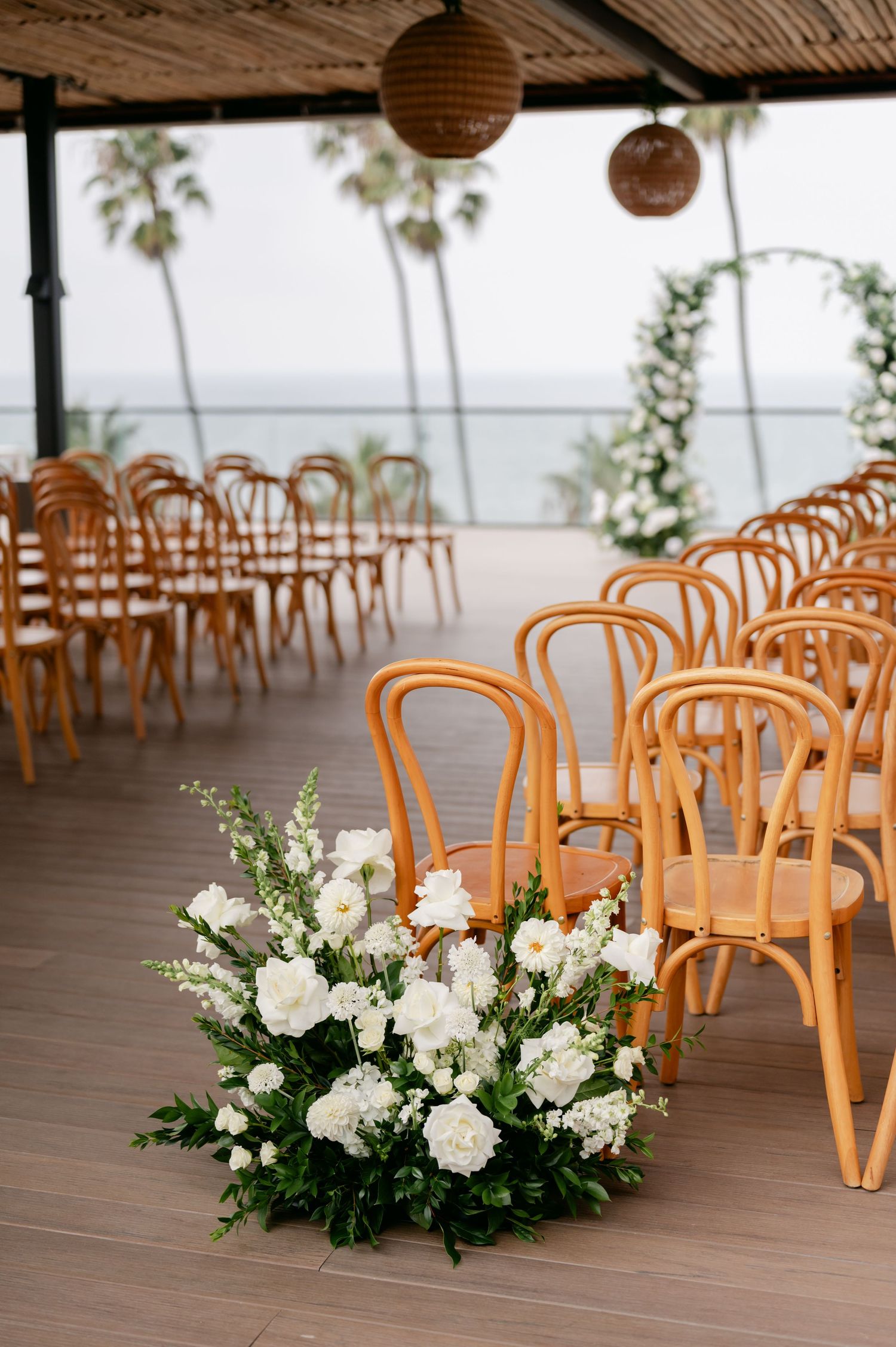 Wooden bentwood chairs and white floral arrangements line a covered outdoor wedding ceremony space.
