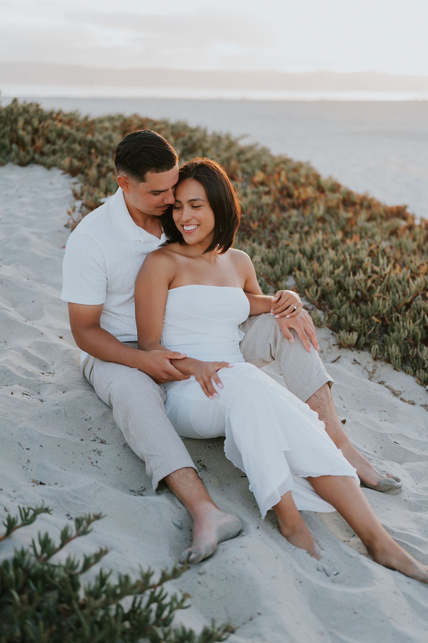 Romantic couple in white outfits embracing on sandy beach during sunset photoshoot.