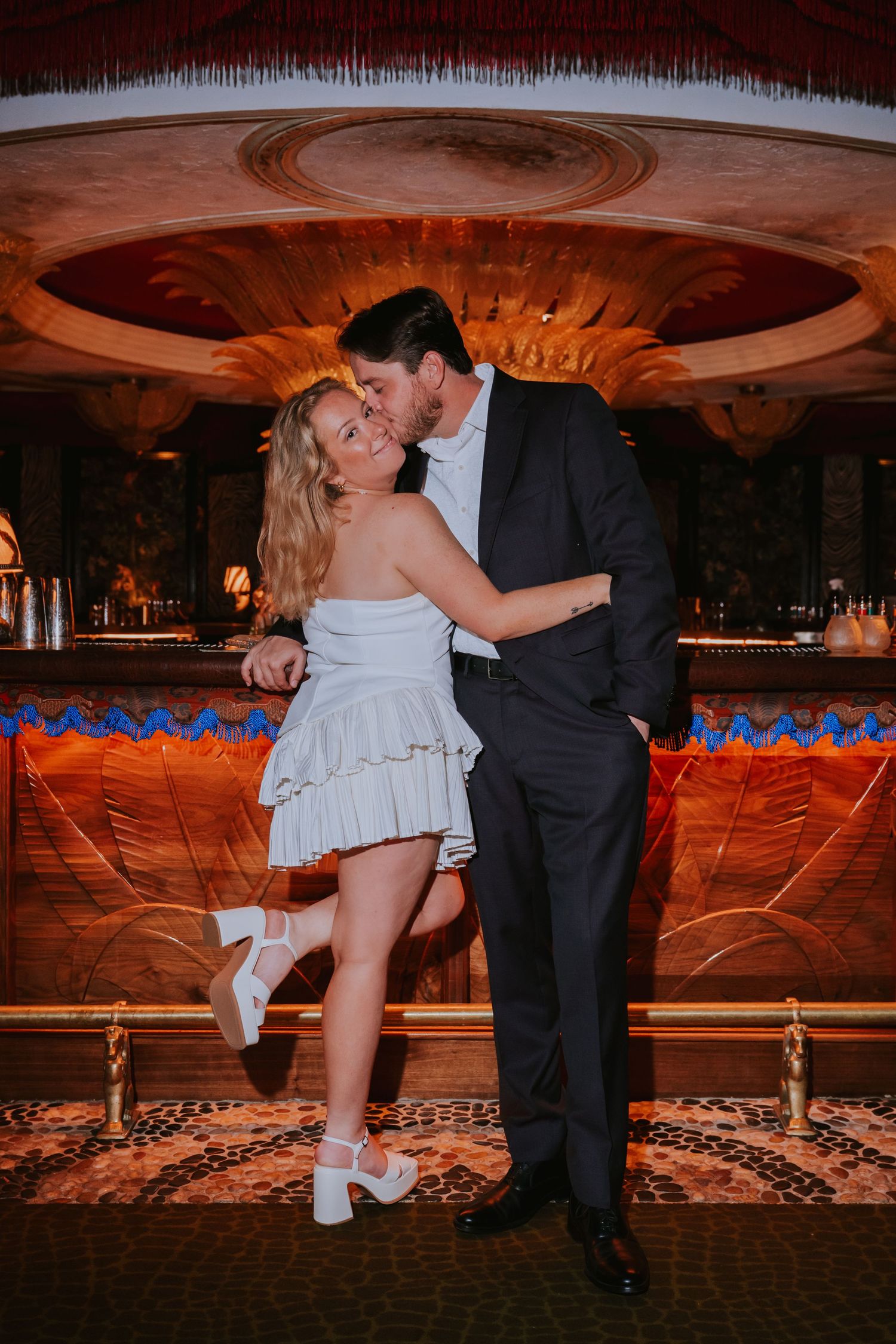 Bride in white ruffled mini dress and groom in dark suit sharing a kiss in a luxurious bar venue.