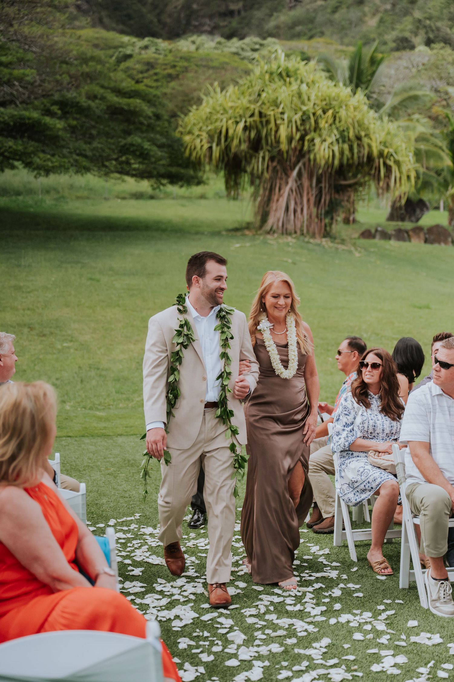 Wedding guests seated on white chairs with flower petals scattered on grass lawn.