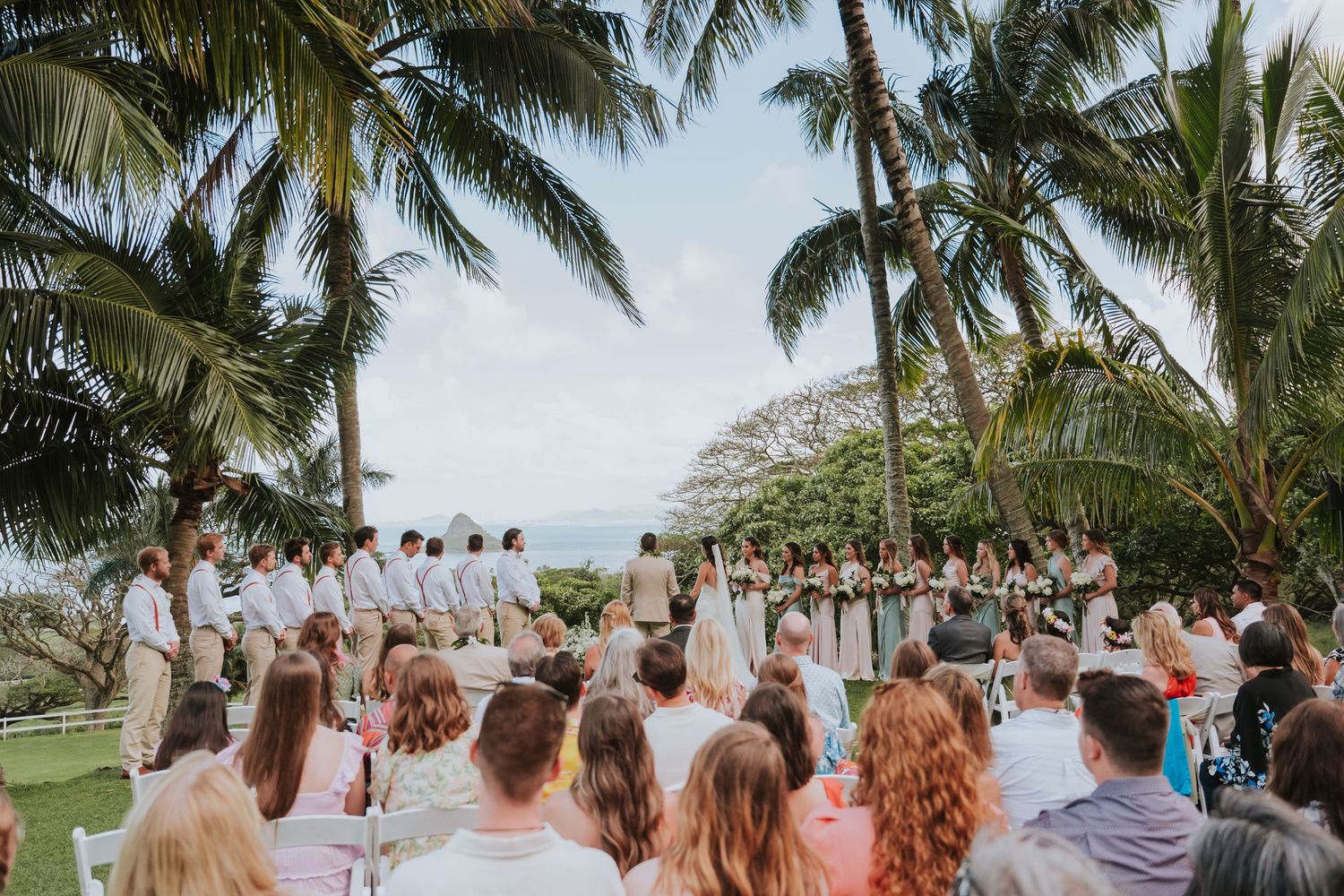 Wedding guests gather under swaying palm trees on a tropical beach during a destination ceremony.