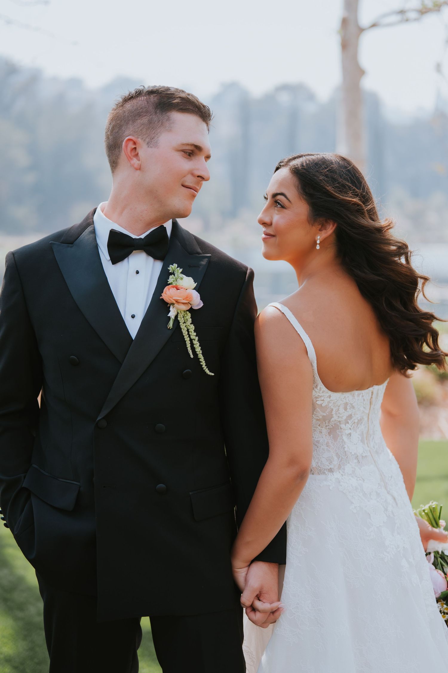 A bride and groom hold hands and gaze at each other, dressed in a white lace gown and black tuxedo outdoors.