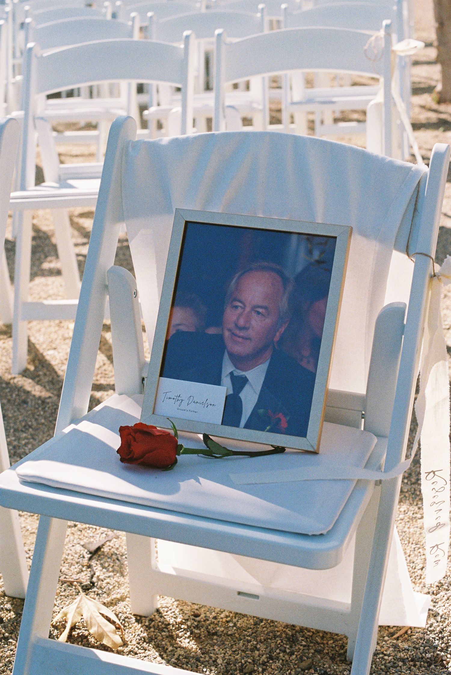 A framed memorial photo with a red rose on a white folding chair at an outdoor wedding ceremony.