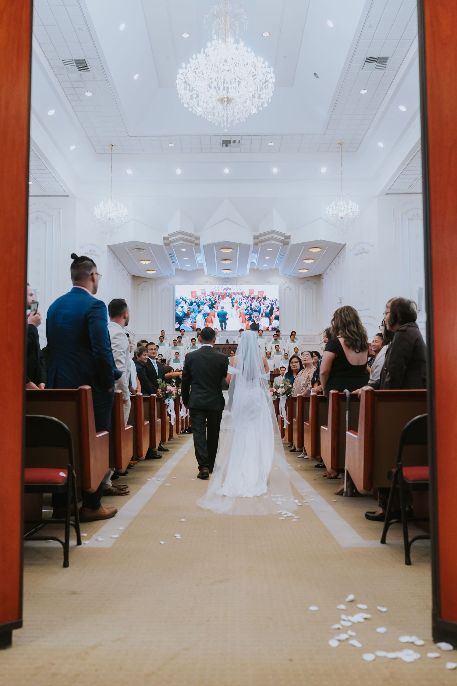 Wide angle view of a wedding ceremony in a bright white ballroom with crystal chandeliers and wooden church pews.