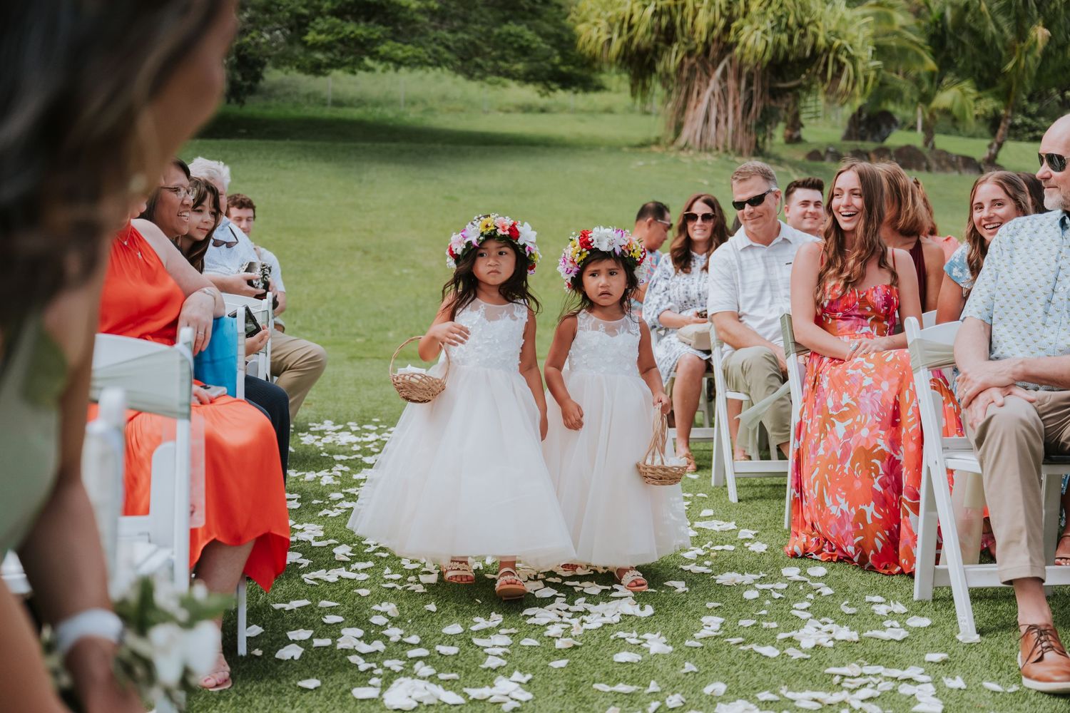 Wedding party members in coral and white dresses stand together on a grassy lawn with scattered flower petals.