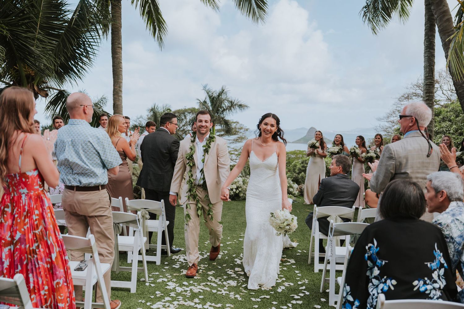 Wedding guests gather under swaying palm trees at a tropical outdoor ceremony venue with white chairs.