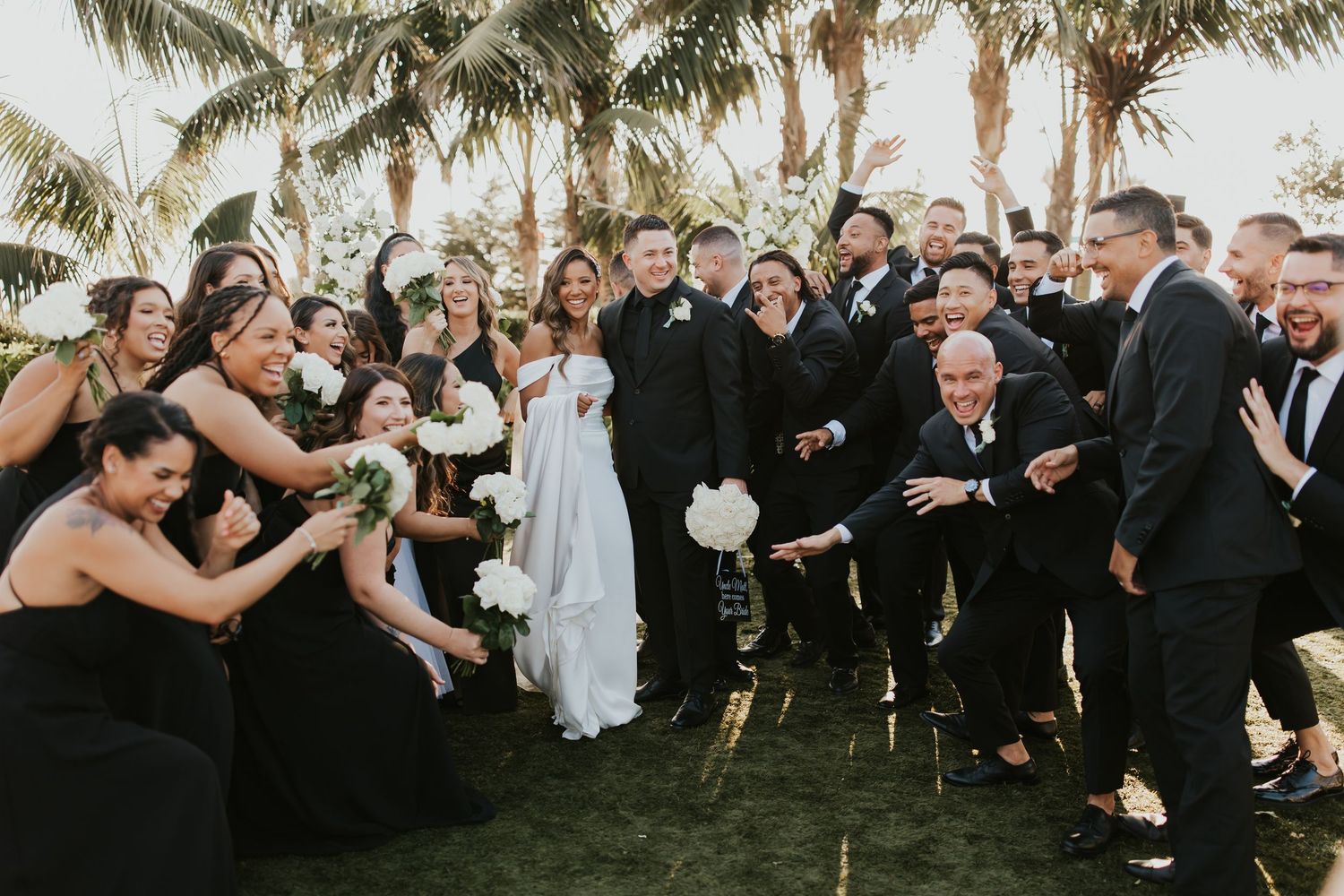 A joyful wedding party poses together outdoors in formal black and white attire for group photos.